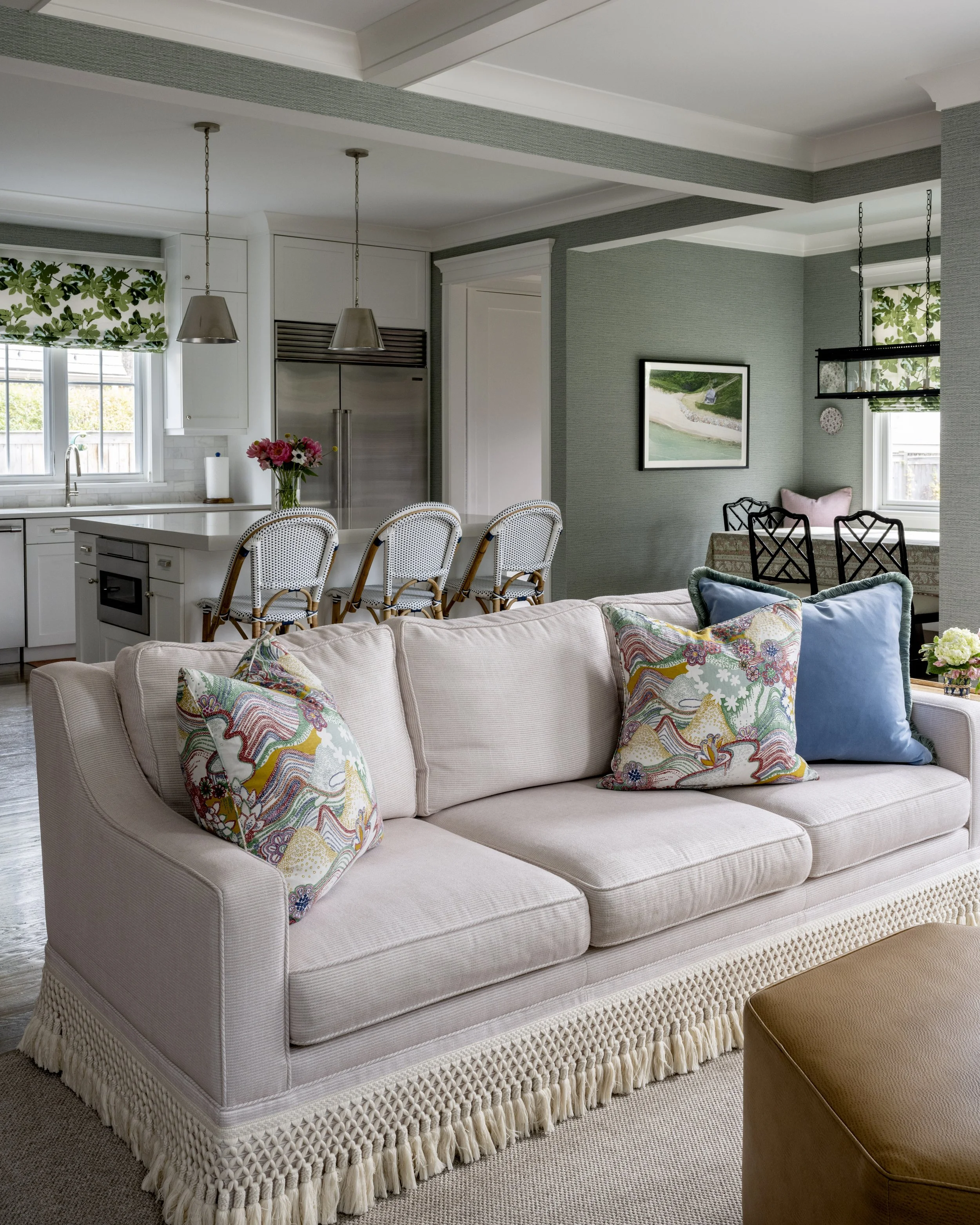 A living room with a beige sofa with colorful patterned pillows, next to a brown ottoman, with a kitchen and dining area in the background.