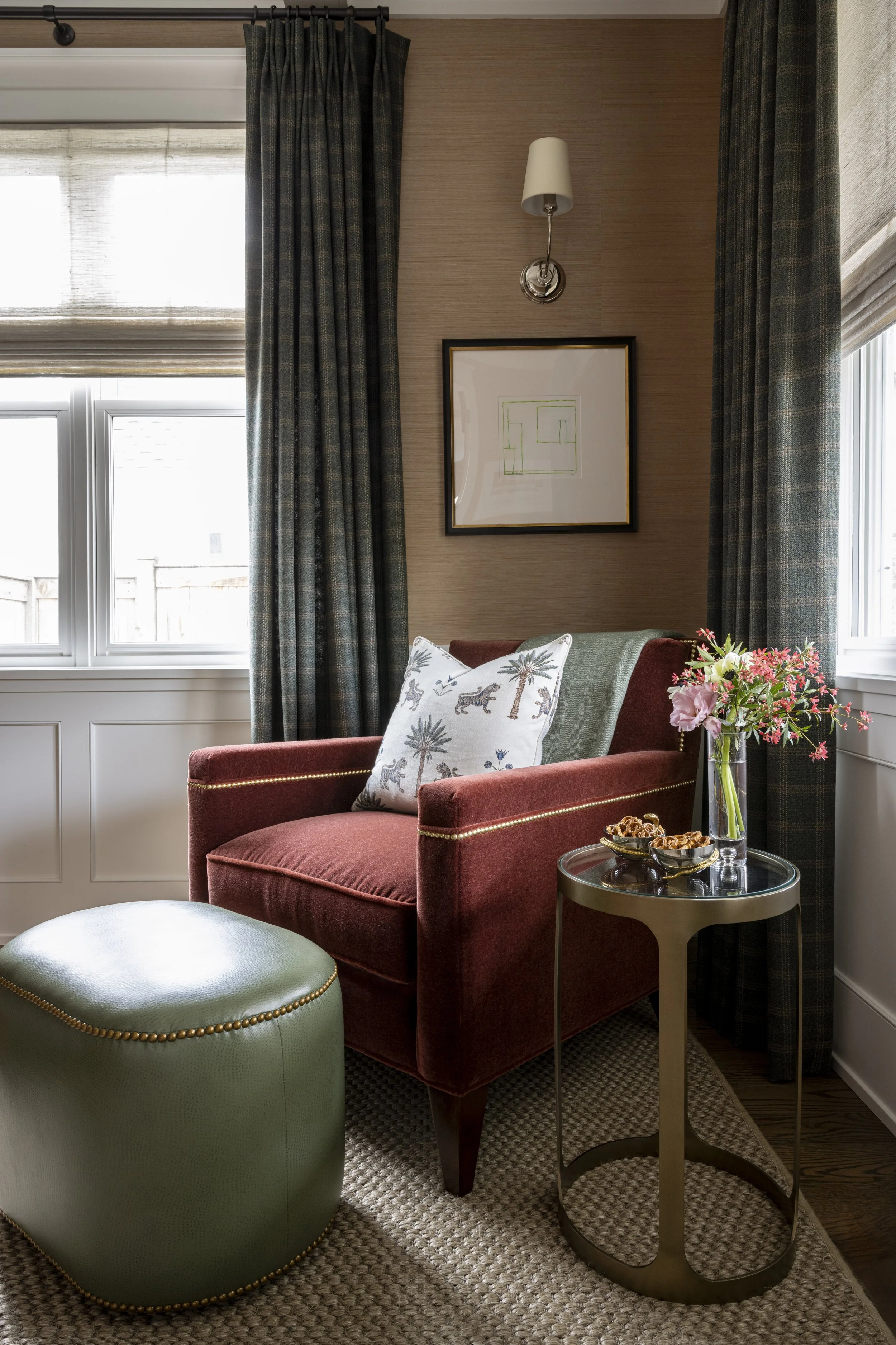 A cozy corner with a red armchair, green ottoman, side table with a vase of pink and white flowers, and framed artwork on a tan wall, next to windows with plaid curtains.