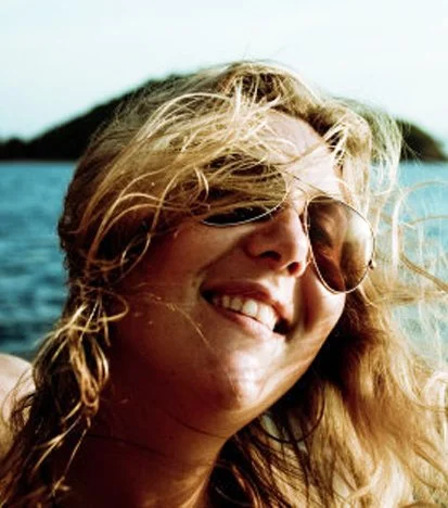Woman with blonde hair and sunglasses smiling at the camera, outdoors near water.