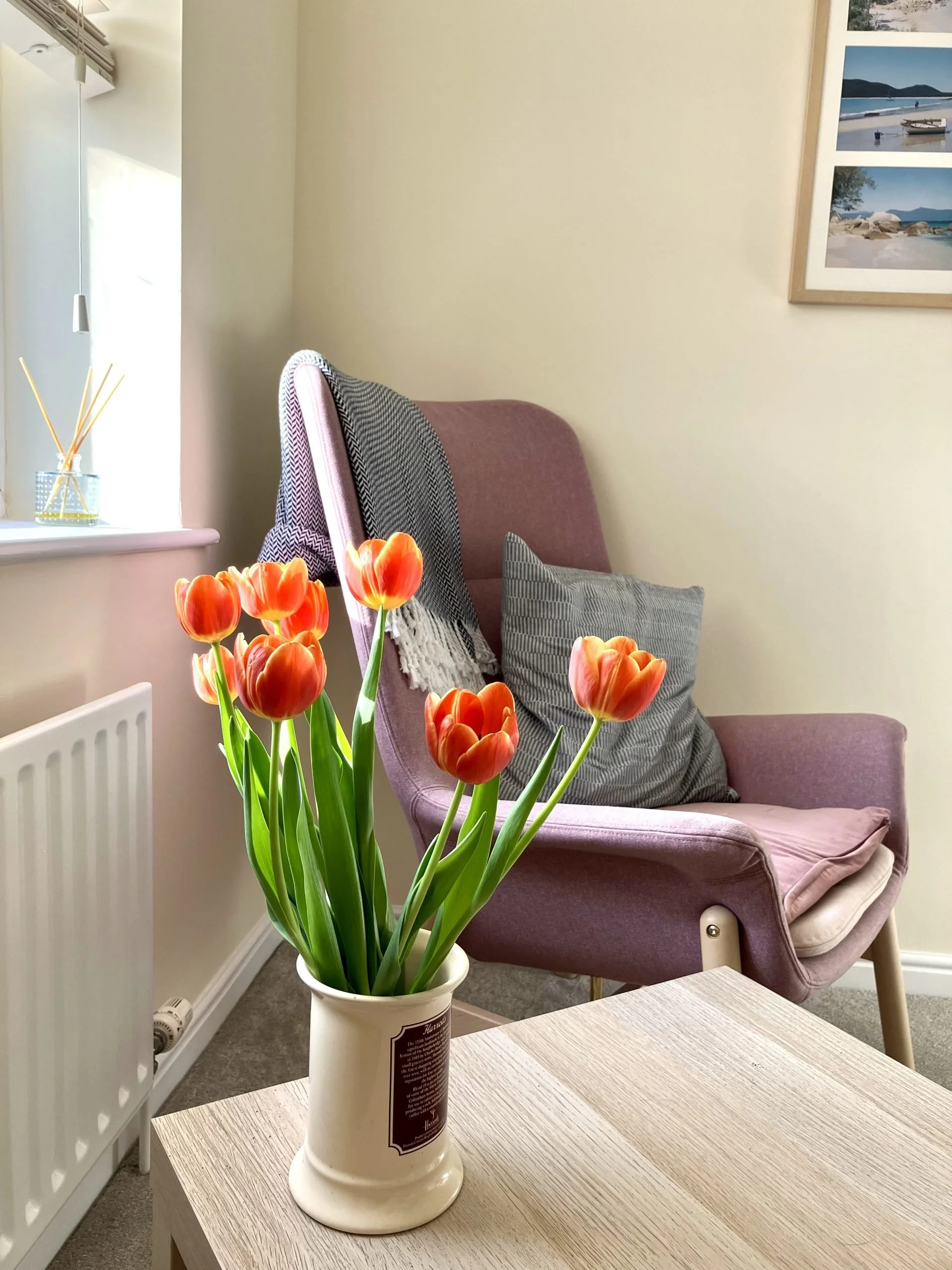A white ceramic vase with orange tulips on a wooden table in a living room with a pink armchair, gray pillow, and framed pictures of boats and landscapes on the wall.