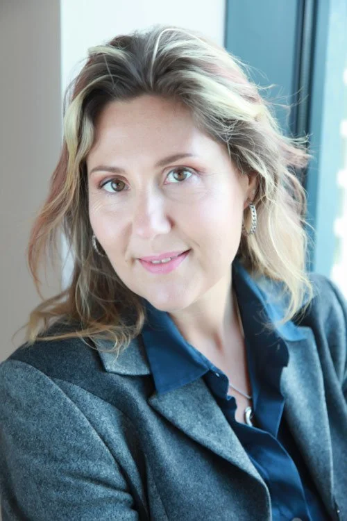 A woman with shoulder-length blond hair and light-colored eyes, wearing a dark blue shirt and gray blazer, smiling while looking at the camera, with a window in the background.