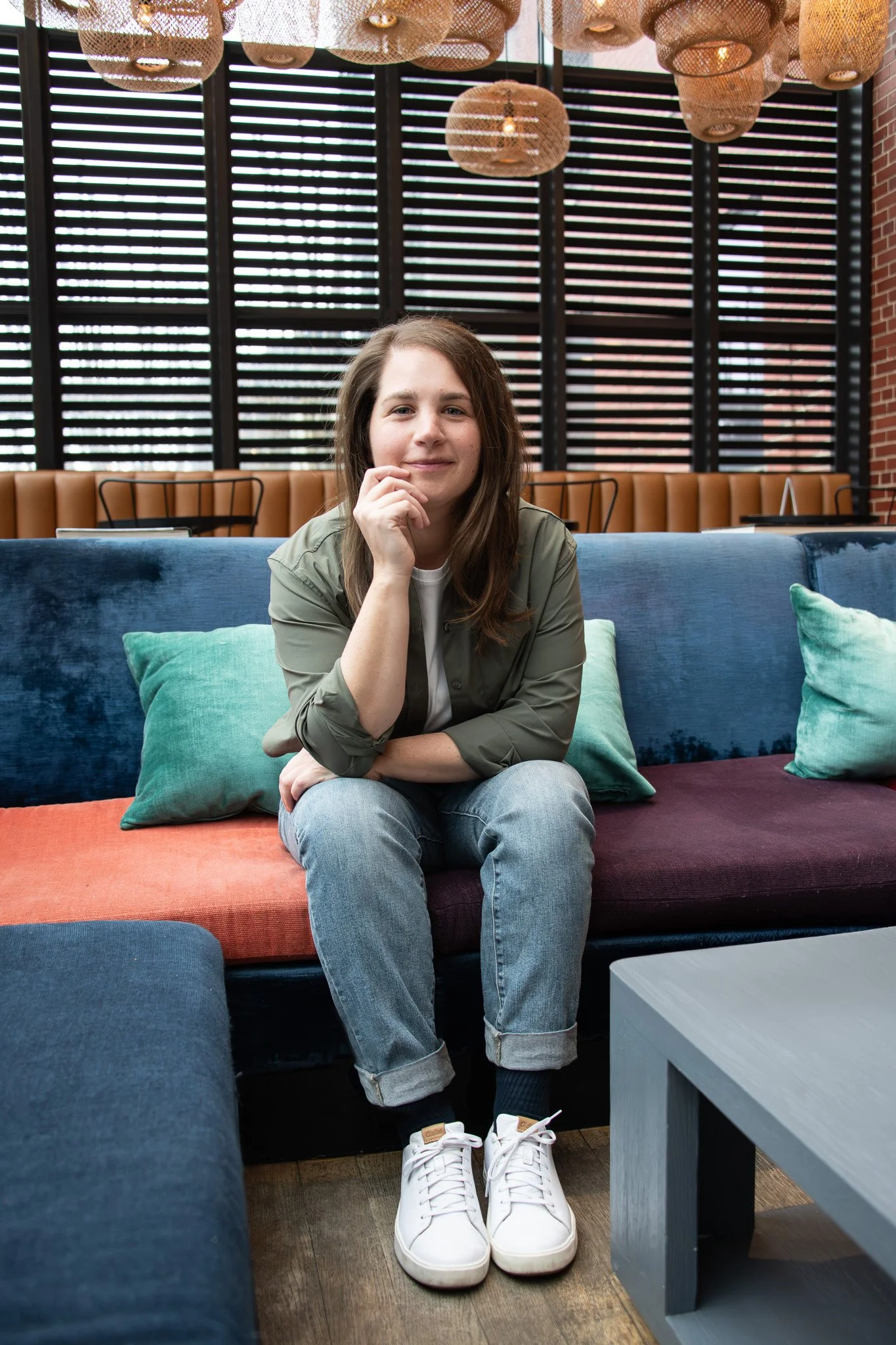 A young woman with long brown hair sitting on a colorful sofa in a modern, stylish lounge or cafe. She is wearing a green jacket, light blue jeans, and white sneakers, with a relaxed and confident expression.
