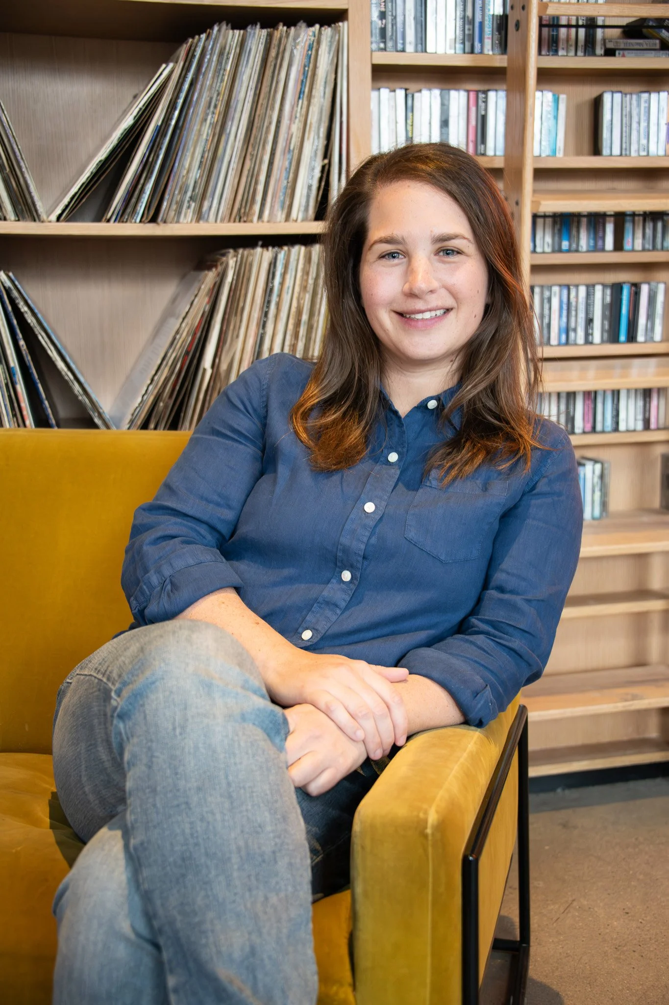 A young woman with brown hair and a blue button-up shirt sitting on a yellow vintage sofa with her hands folded, smiling, against a background of wooden bookshelves filled with vinyl records and CDs.