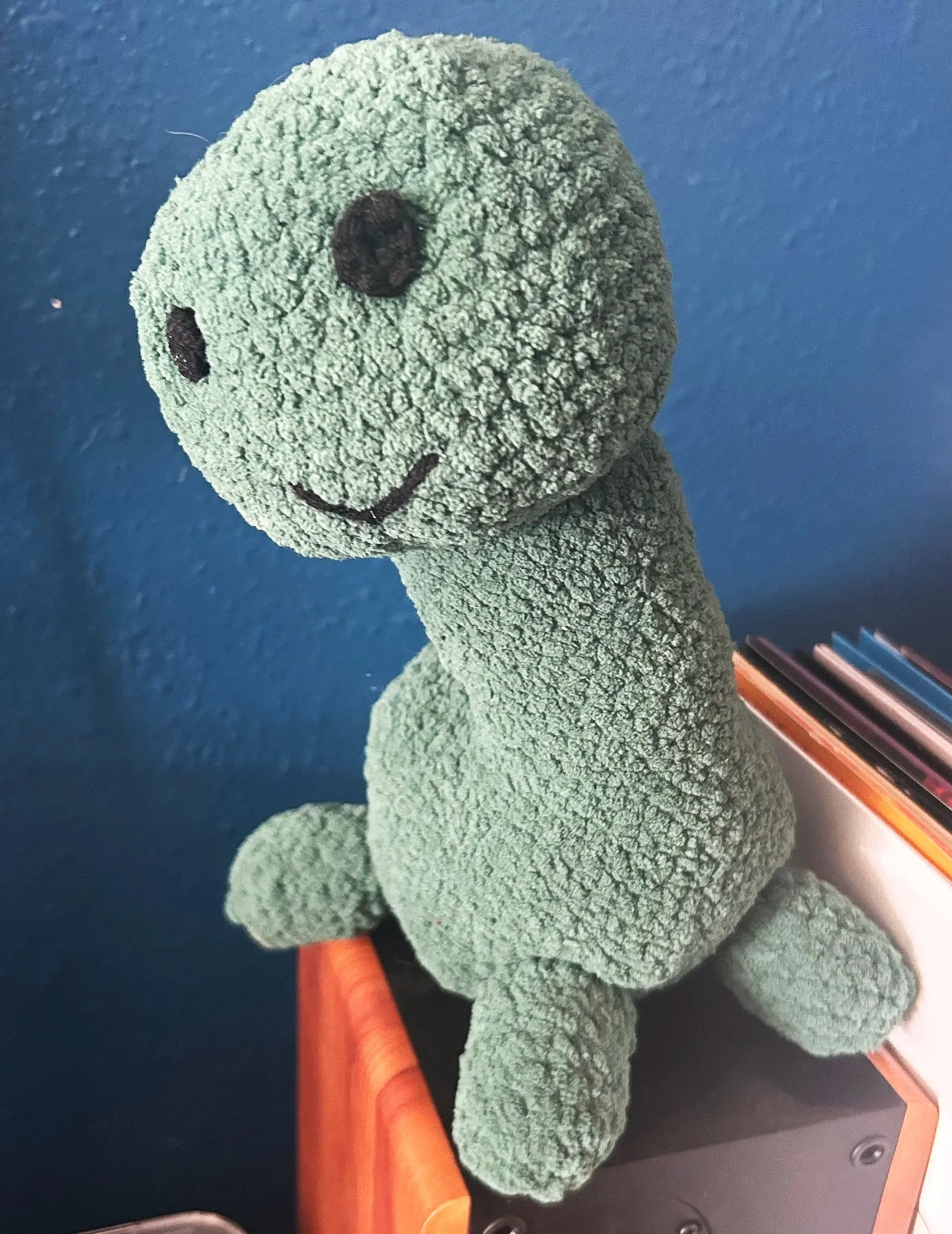 A plush green dinosaur toy with a smiling face, sitting on a wooden surface against a blue wall.