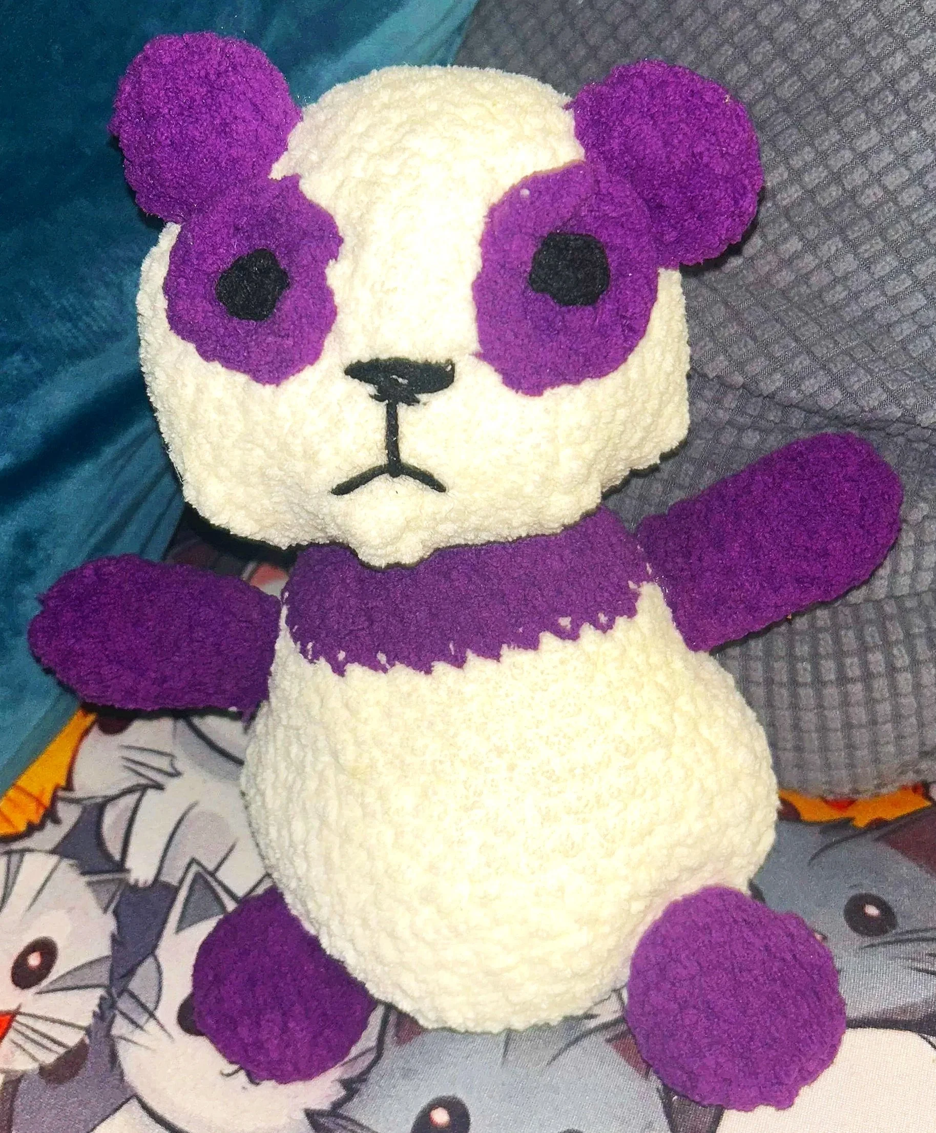 A plush panda bear toy with purple ears, eye patches, arms, and feet, and a purple band around its neck, sitting on a surface with cartoon cats and mice.