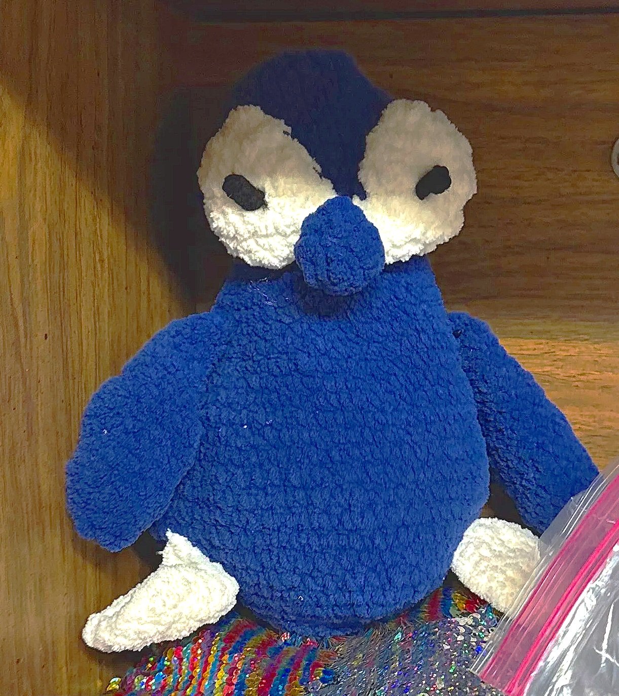 A plush penguin, with a blue body, white face, and black eyes, sitting on a colorful, textured surface.