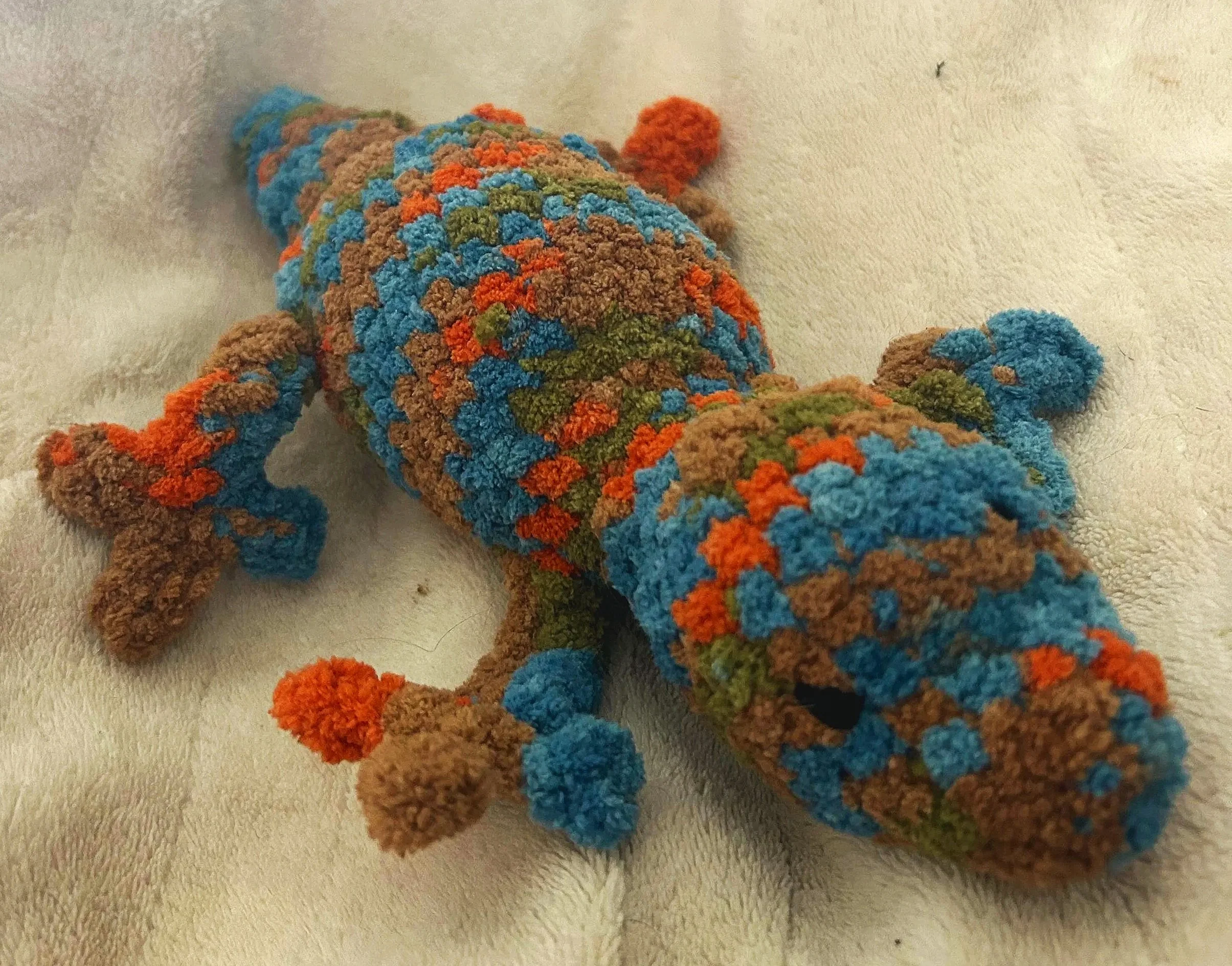 Colorful handmade crocheted lizard toy with blue, orange, green, and brown patches, lying on a light-colored fabric surface.
