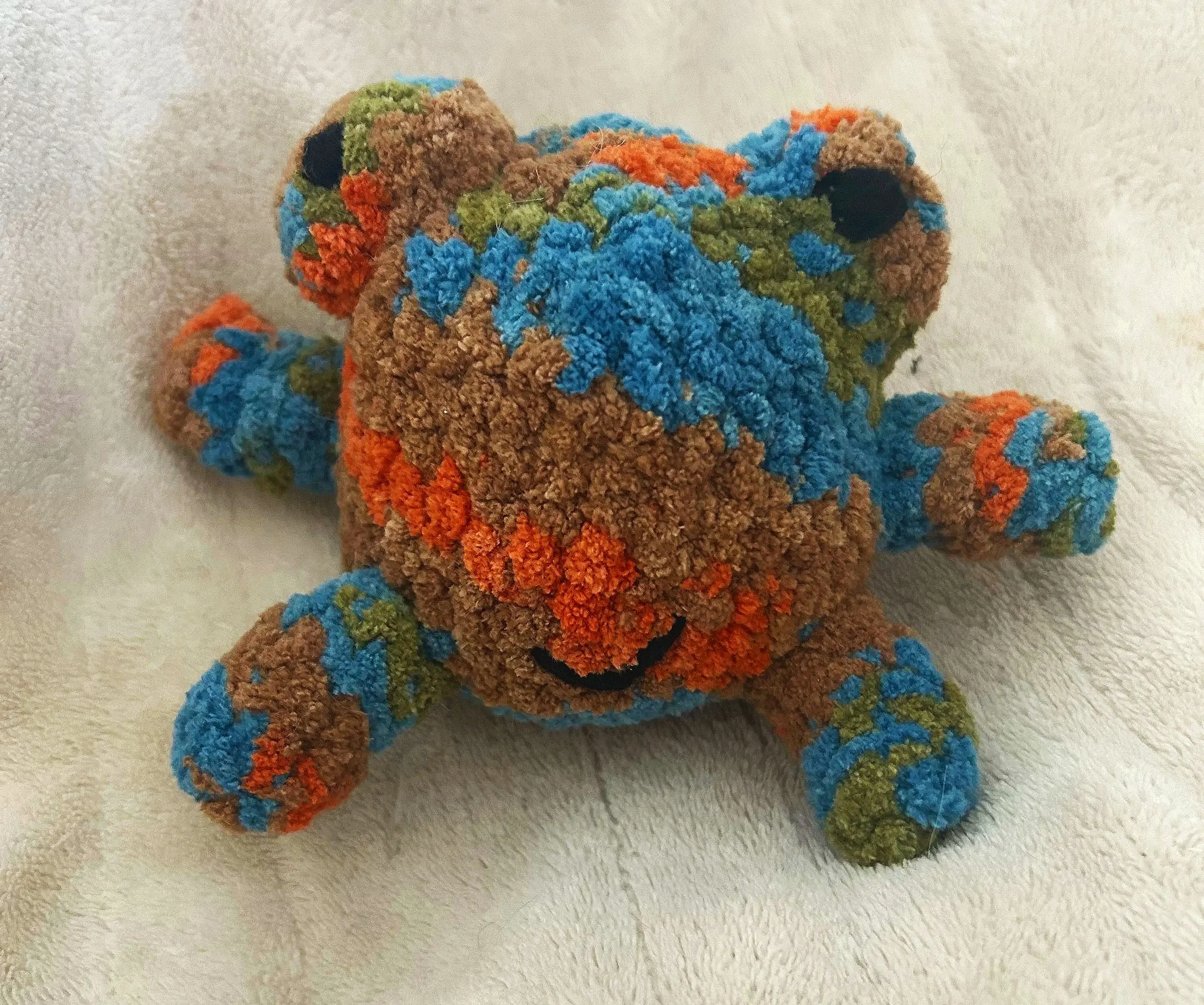 Colorful plush toy frog with brown, blue, green, and orange patches lying on a light-colored surface.