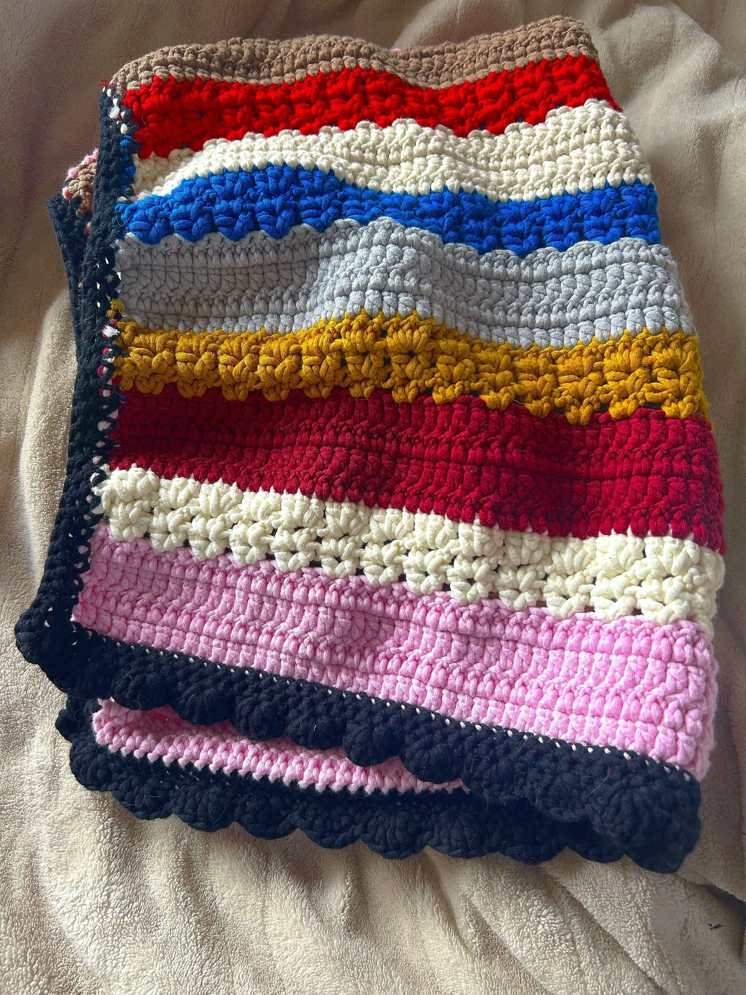 A folded, colorful hand-crocheted blanket with a scalloped black border, placed on a cream-colored surface.