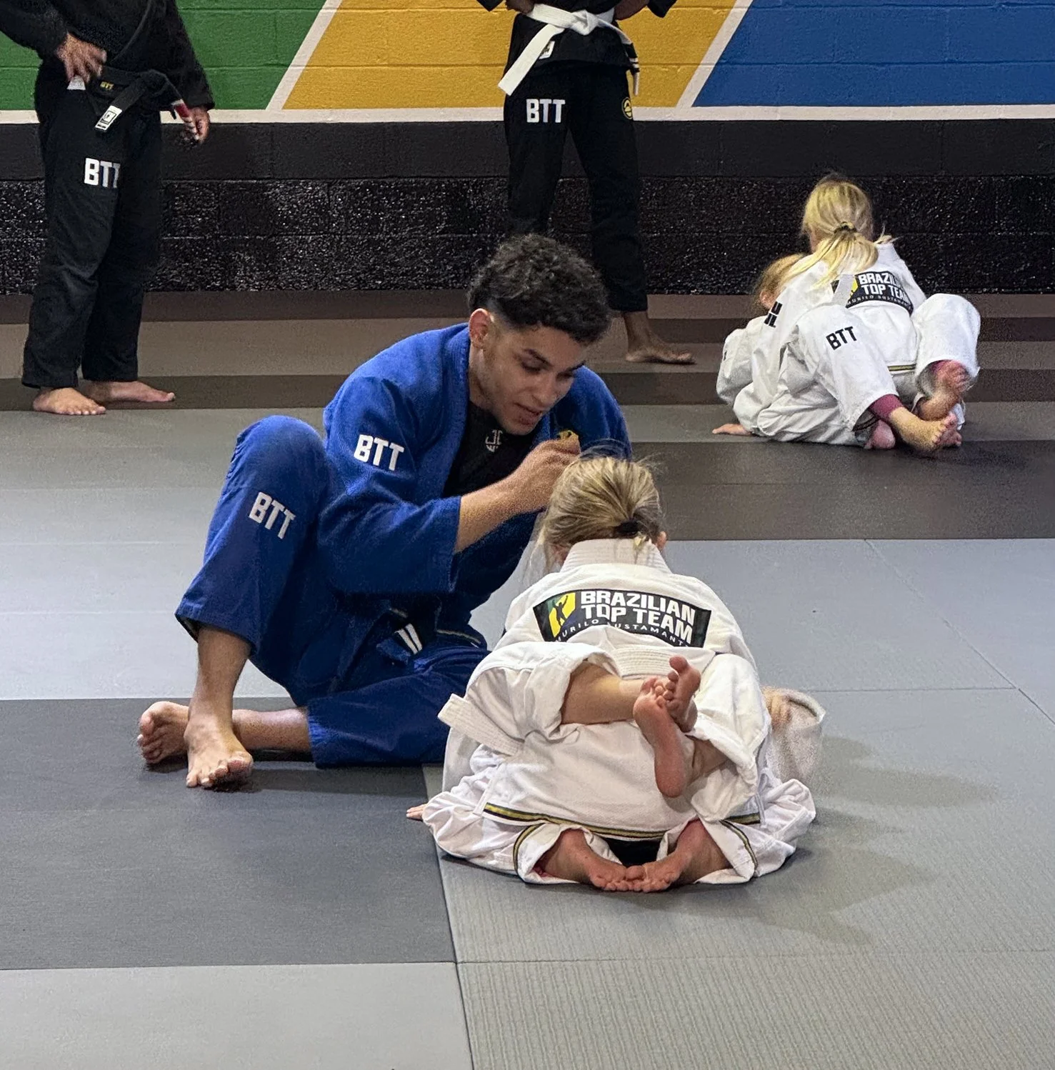 Brazilian Jiu-Jitsu class with young children practicing techniques, one child in white belt on ground and instructor in blue gi assisting, others in background practicing.