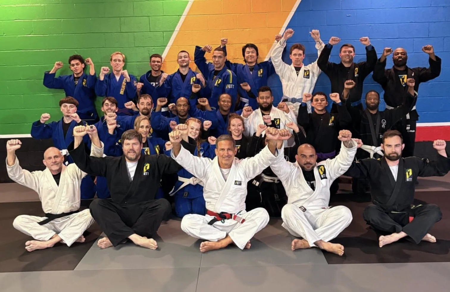 Group of mixed gender Brazilian Jiu-Jitsu practitioners in gi uniforms, posing with fists raised in victory inside a gym with colorful walls.