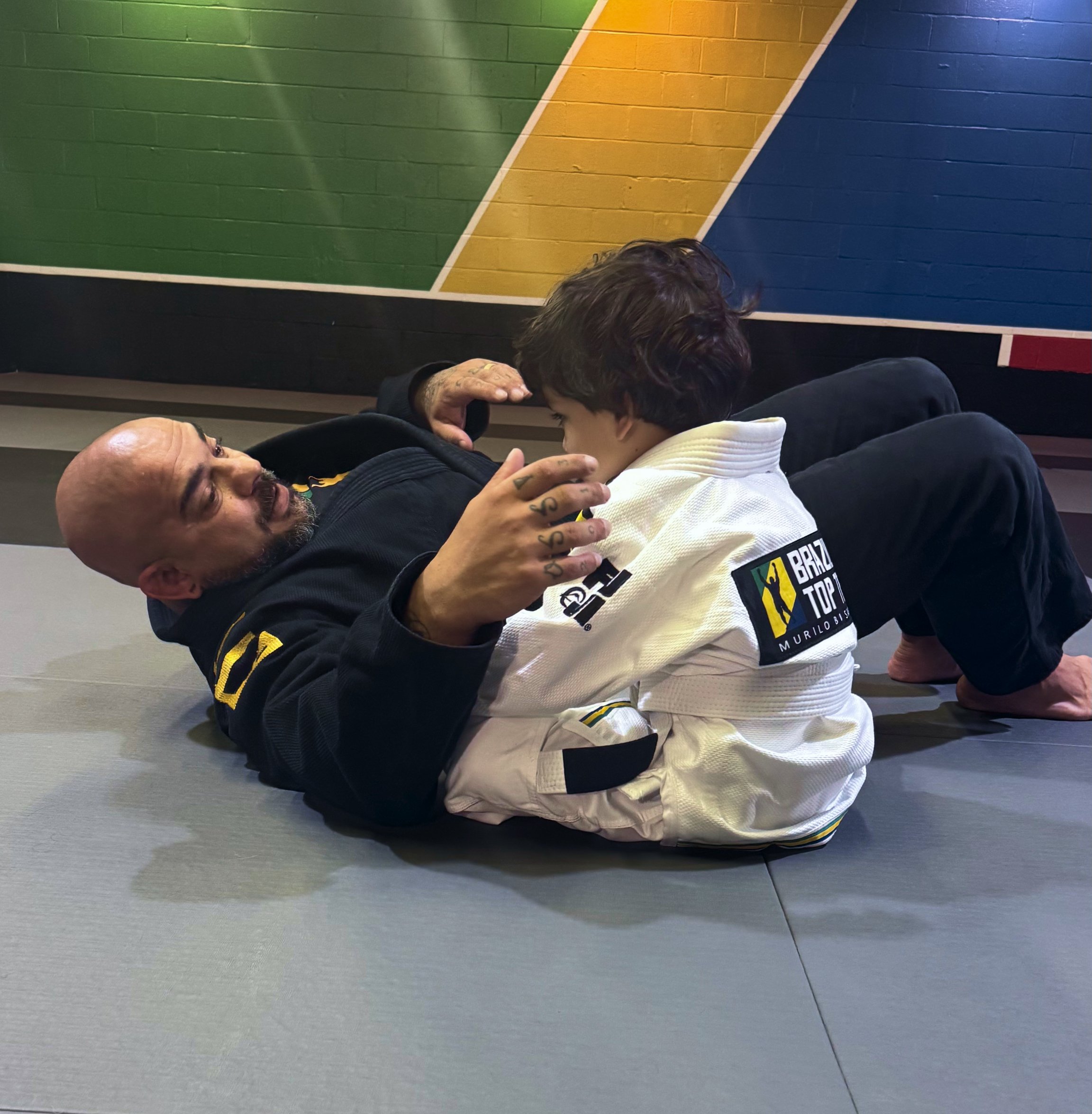 A man lying on the floor with a child, possibly practicing Brazilian Jiu-Jitsu. The man has a bald head and tattoos on his hand. The child is wearing a white gi with patches, and the man is wearing a black gi. They are in a martial arts gym, with a decorated wall in the background.