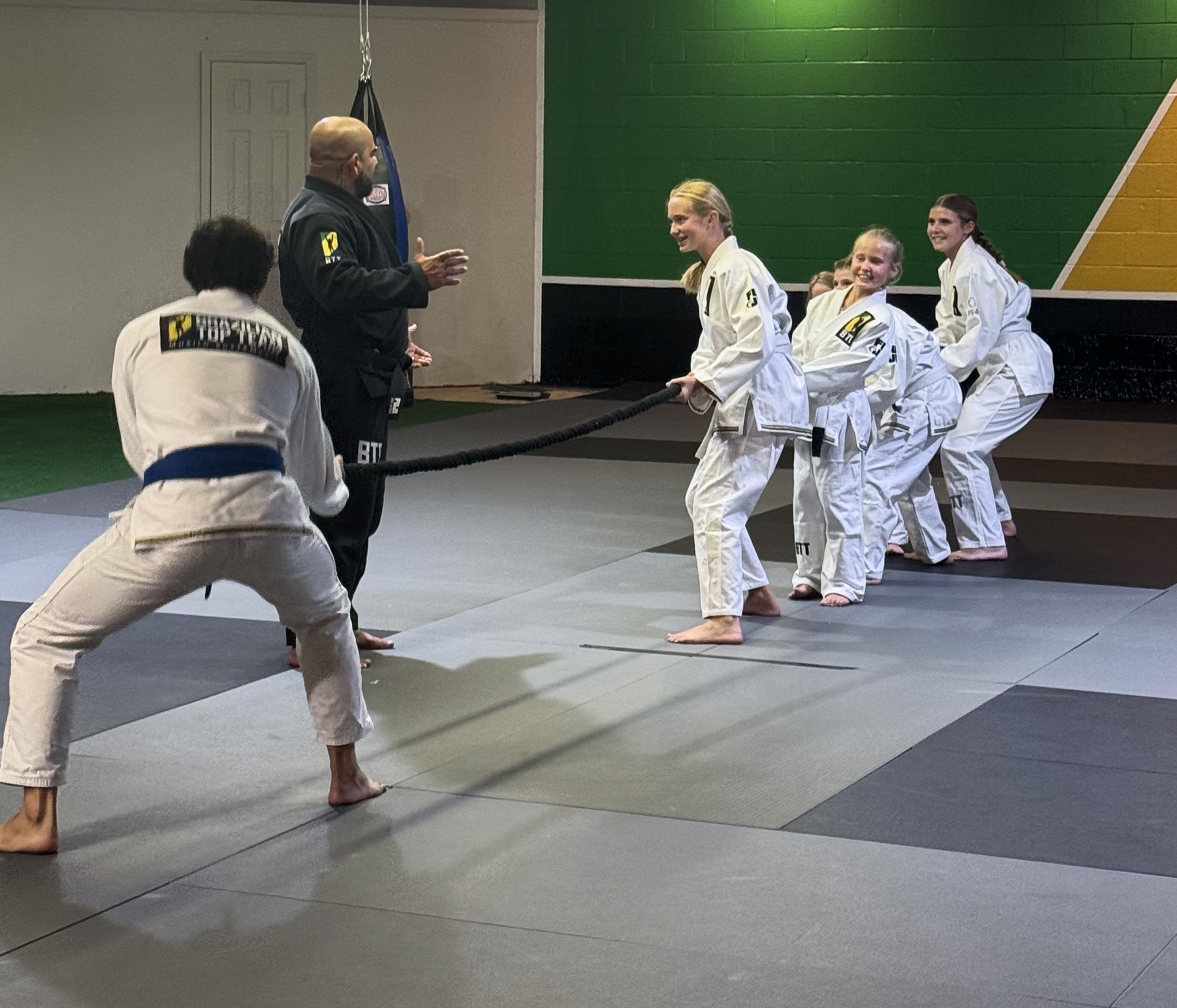 Group of young girls in gi uniforms participating in a martial arts class, some smiling, with an instructor talking to them in a gym with green and yellow wall panels.