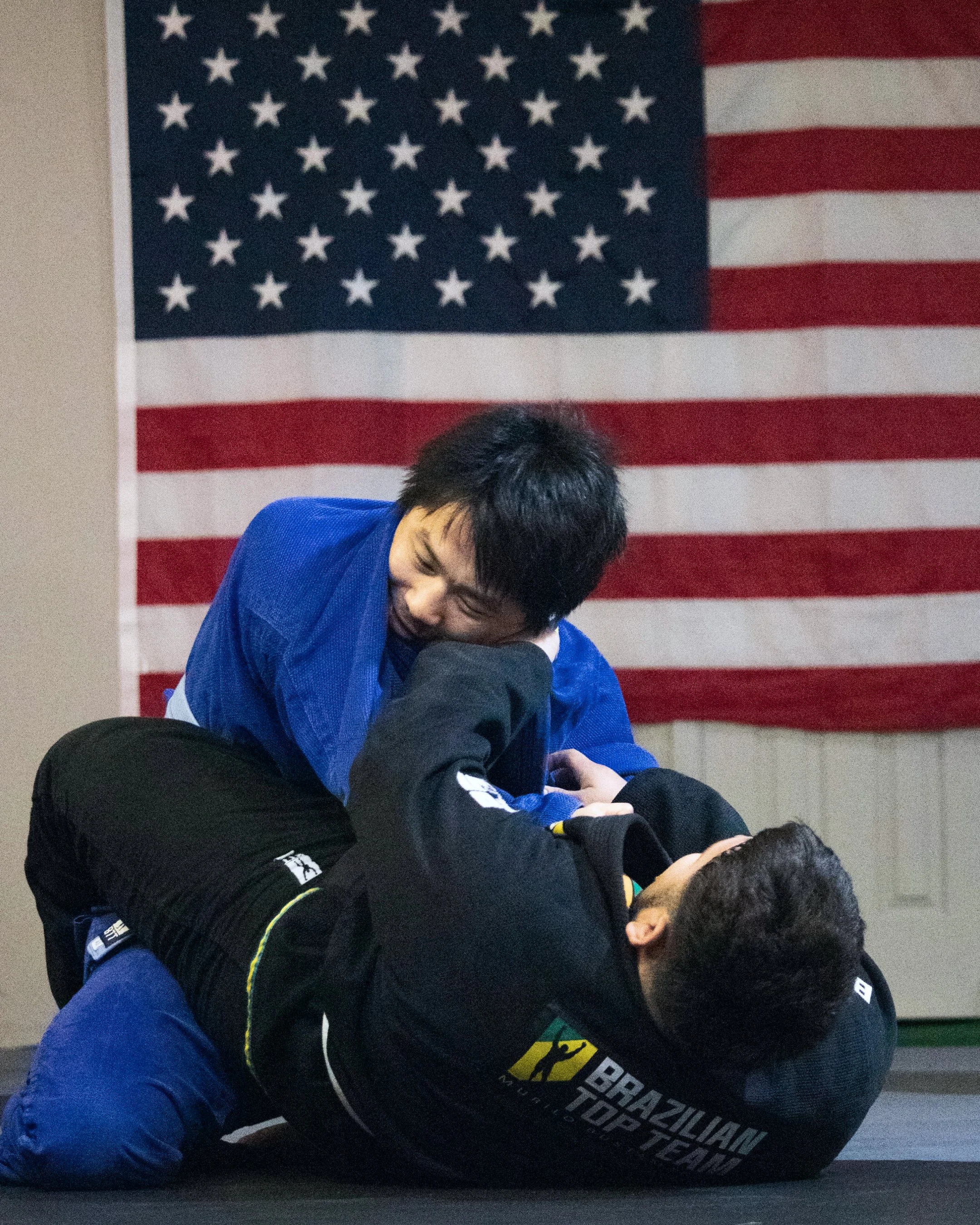 Martial arts practitioners training on mats in front of American flag backdrop, wearing uniforms with patches and logos.