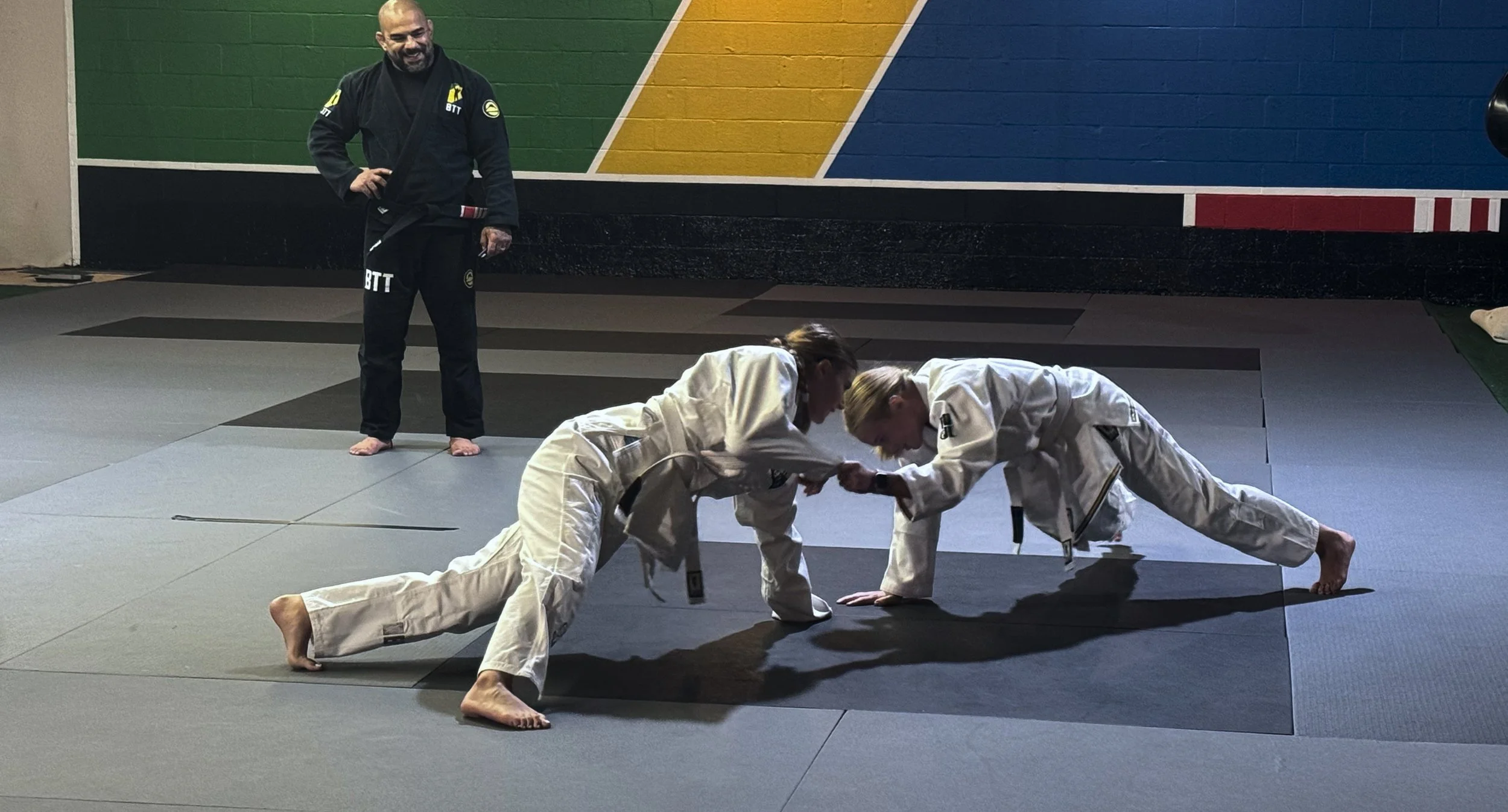 Two women practicing Brazilian Jiu-Jitsu on a gray mat while a coach watches. Both women are in white gis, and one is attempting to control the other. The coach stands in the background against a colorful painted wall.