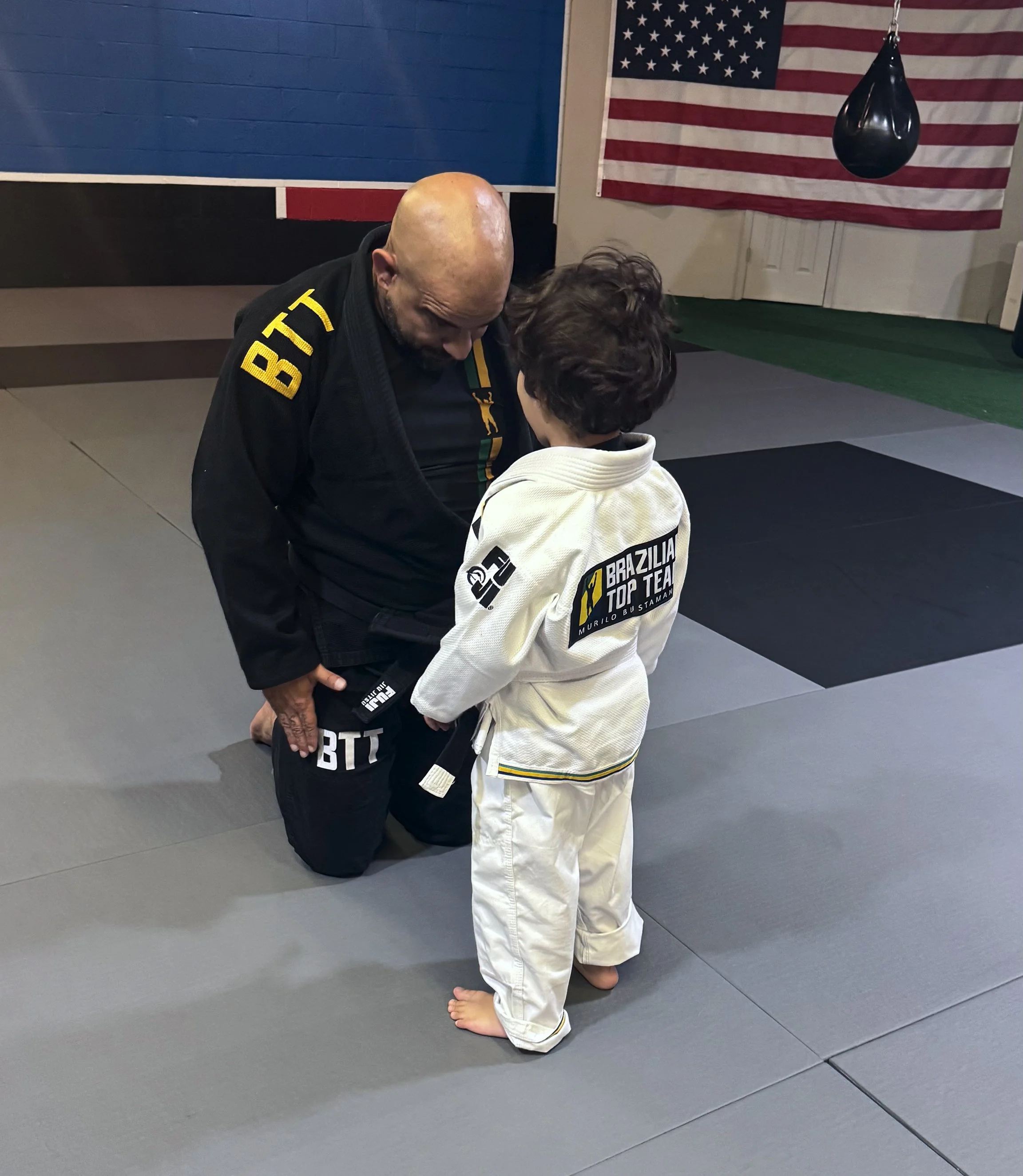 A bald man and a young boy are practicing Brazilian Jiu-Jitsu on a gray mat in a martial arts gym. The man is wearing a black gi with yellow patches, and the boy is wearing a white gi with patches. The man is kneeling and looking down at the boy, who is standing with his head tilted downward. In the background, there is an American flag hanging on the wall and a black punching bag.