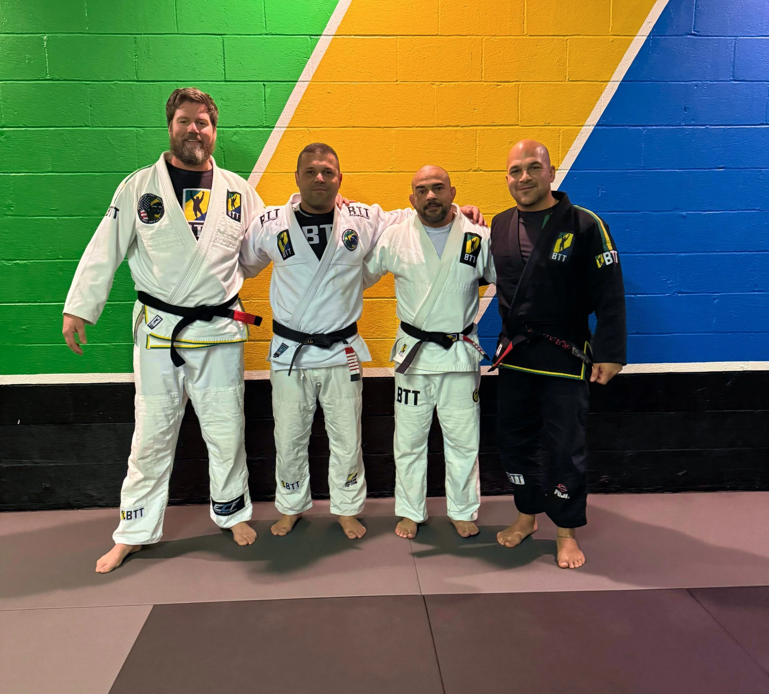 Four men standing on a martial arts mat in front of a colorful wall painted with green, yellow, and blue stripes. Three of them are wearing white jiu-jitsu gis with black belts, and one is wearing a black gi with a black belt. They are posing for the photo with their arms around each other's shoulders.