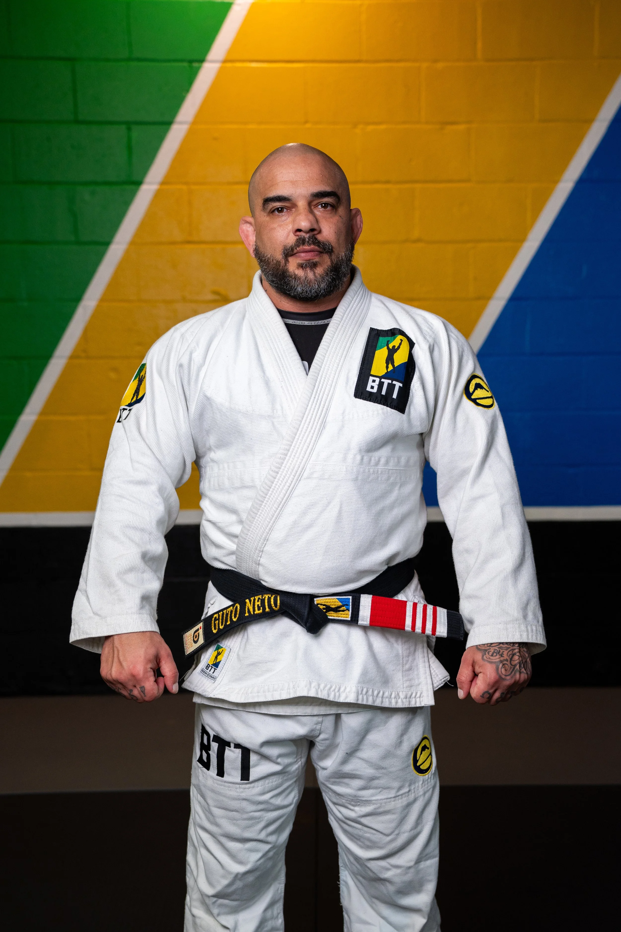 A man in a white Brazilian Jiu-Jitsu gi standing in front of a multicolored background with green, yellow, and blue sections. He has a bald head, beard, and tattoos on his hands. The gi patches indicate martial arts affiliations.