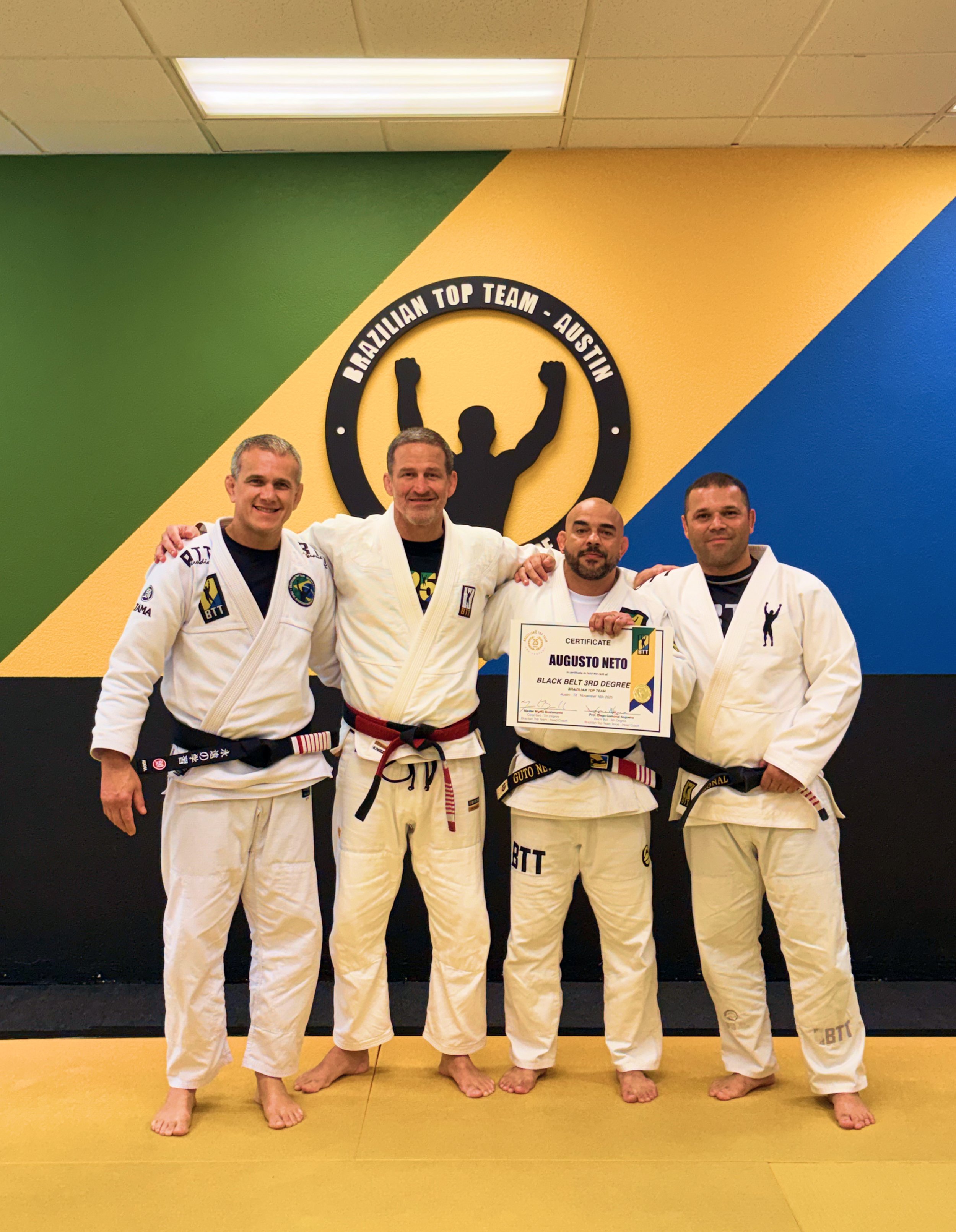 Four men in jiu-jitsu uniforms standing side by side, smiling with arms around each other's shoulders in a martial arts dojo, with a green, yellow, and blue wall and a banner reading 'Brazilian Top Team - Austin' behind them. One man in the center holds a certificate.
