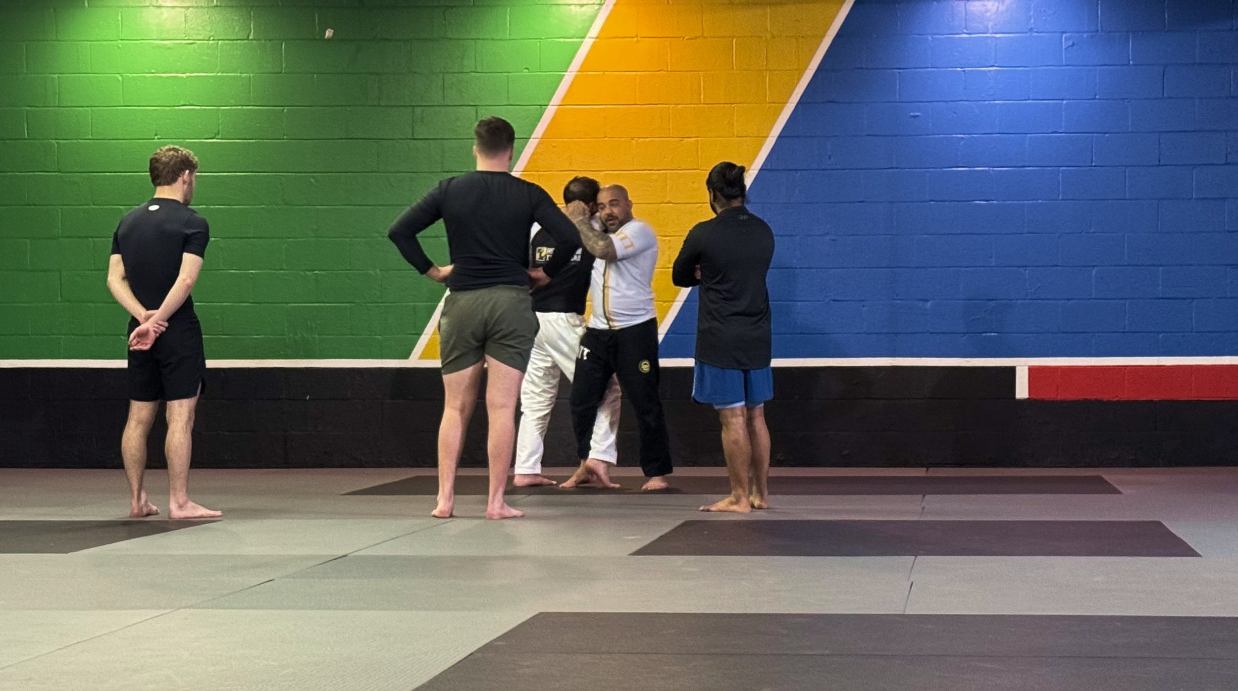 Five men in athletic clothing stand on a yoga mat in a gym with a colorful wall, engaging in a discussion or instruction session.