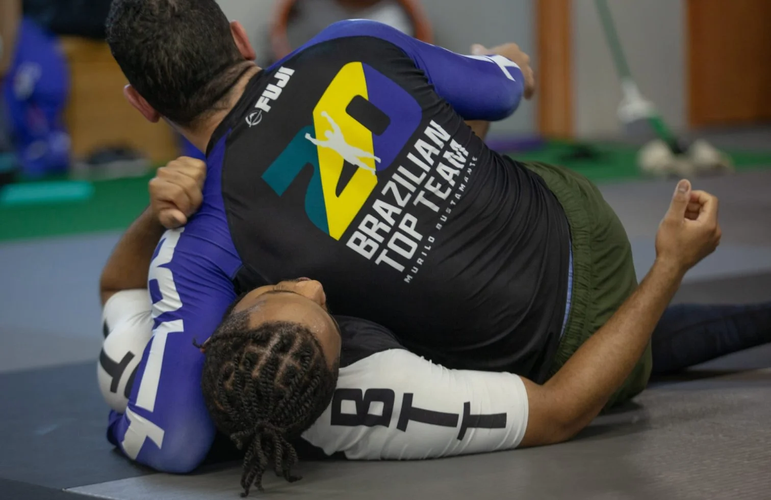 Two Brazilian Jiu-Jitsu competitors grappling on the ground during a match, one in a black gi and the other in a white gi, on a gray mat in an indoor training facility.