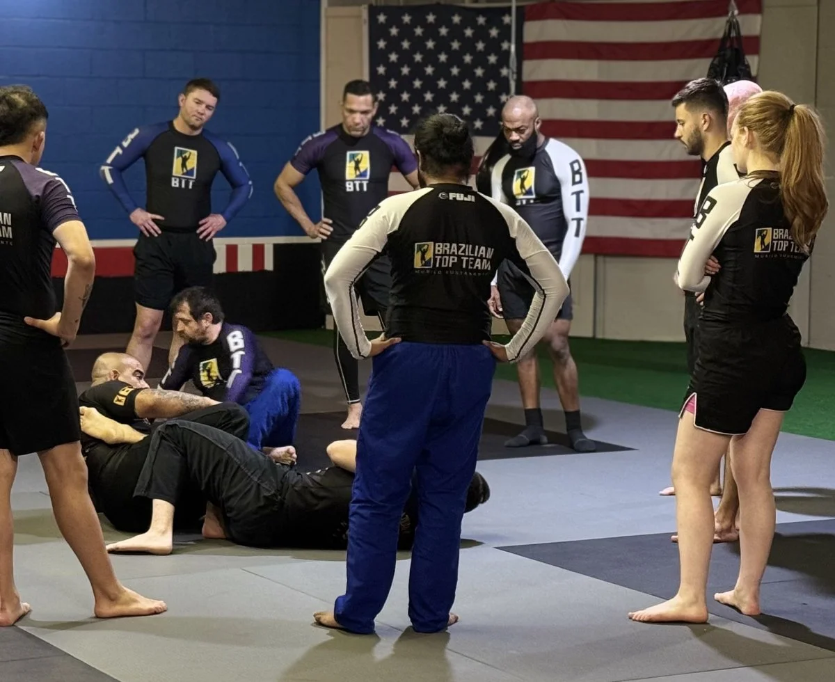 Martial arts students wearing black and white uniforms with 'Brazilian Top Team' logos gather around an instructor who is demonstrating on a person lying on the mat inside training gym, with American flag on the wall.