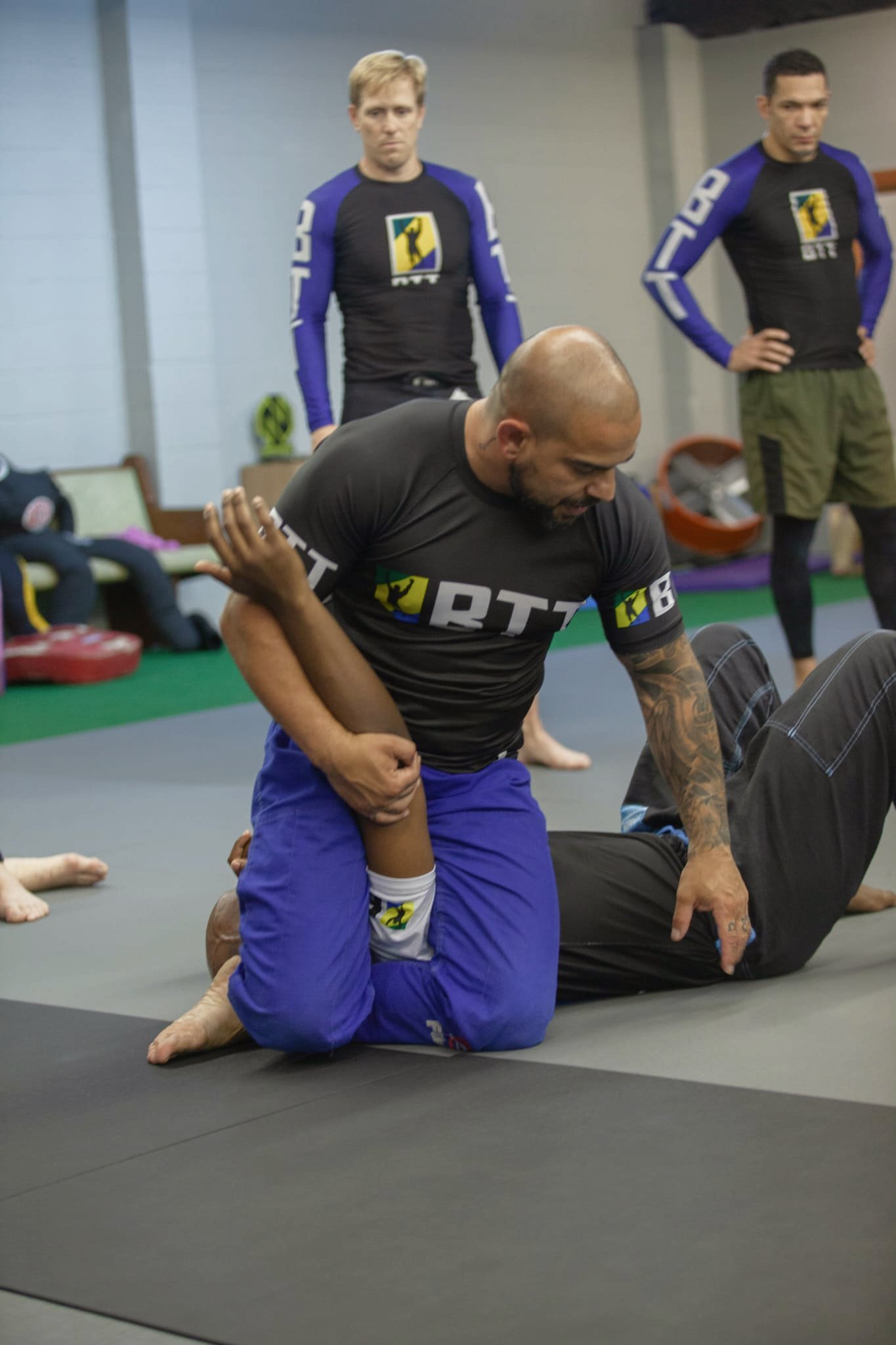 Two people practicing Brazilian Jiu-Jitsu on black mats; one person in blue shorts and a black shirt is on the ground, another in black shorts and a black shirt is kneeling next to him; two men in the background are watching, wearing black shirts with a yellow and black logo and blue accents.