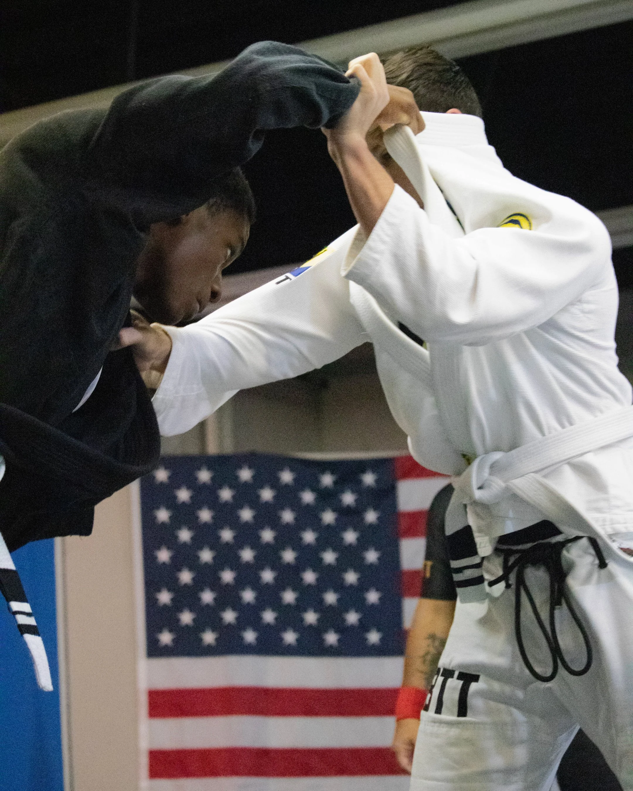 Two young martial artists practicing hand fighting in front of an American flag. One is wearing a black gi with a black belt, and the other is wearing a white gi with a black belt.