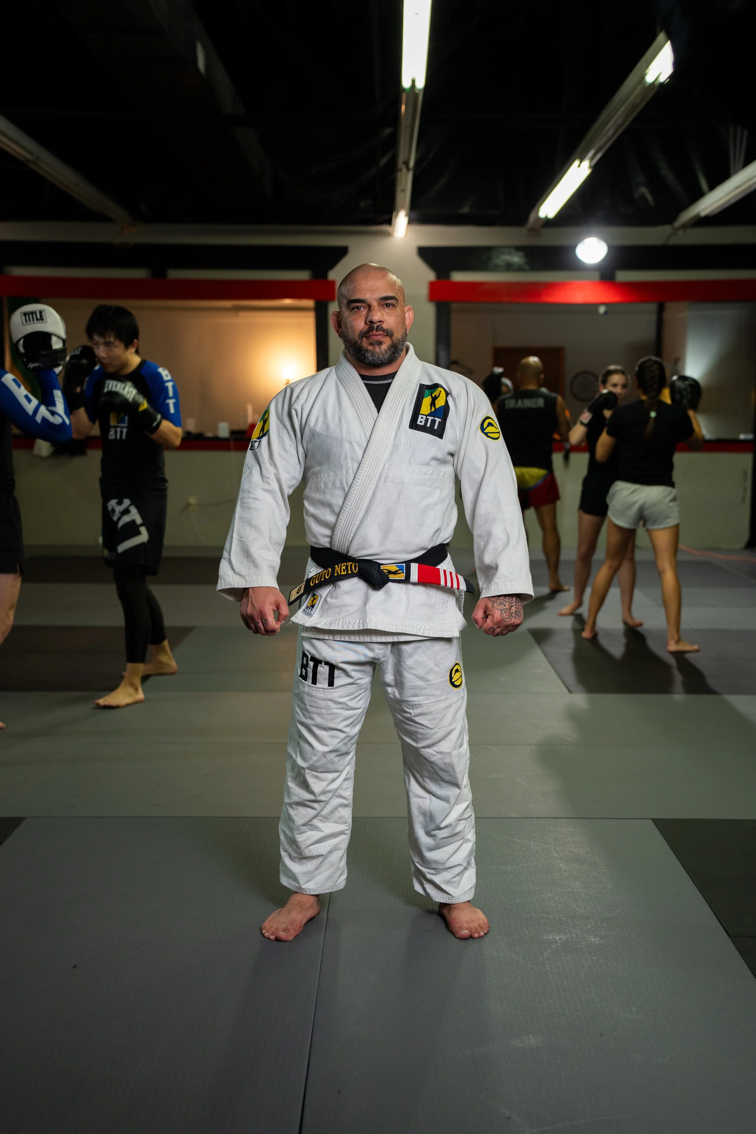 A man standing in a martial arts uniform with a black belt in a martial arts training facility, with other people training in the background.