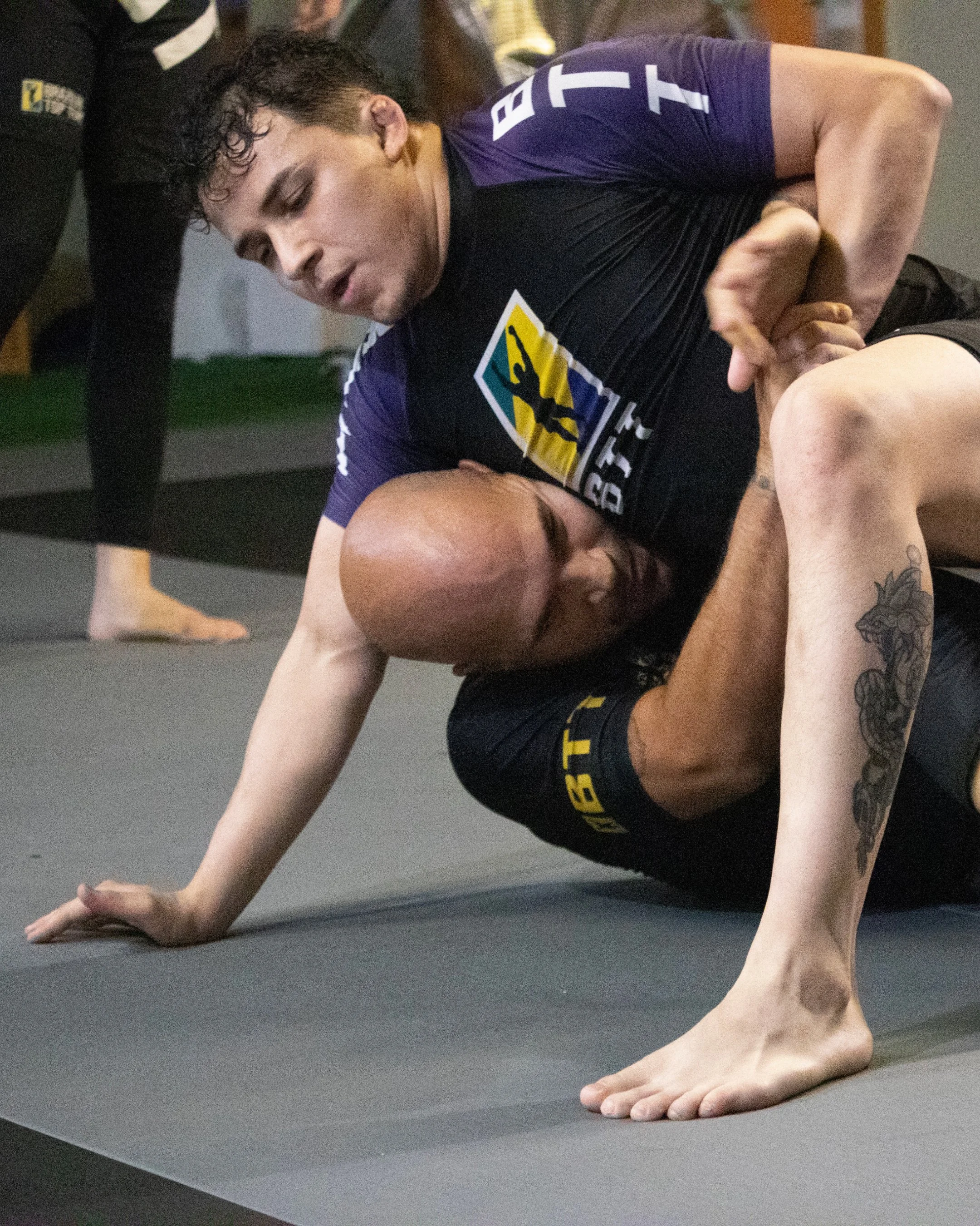A man with curly dark hair practicing yoga on a black mat, in a plank position with one arm extended forward and the other resting on the floor. He is wearing athletic clothing with a logo on his shirt and shorts.