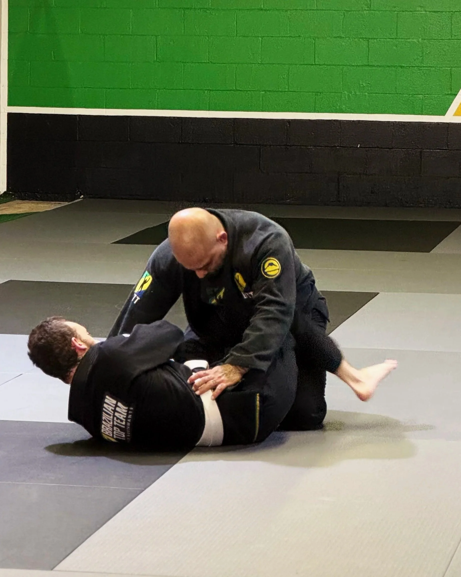 Two men practicing Brazilian jiu-jitsu on the mats in a gym. One man is applying a chokehold on the other, who is lying on his back. The gym has a green and black wall with a white border.