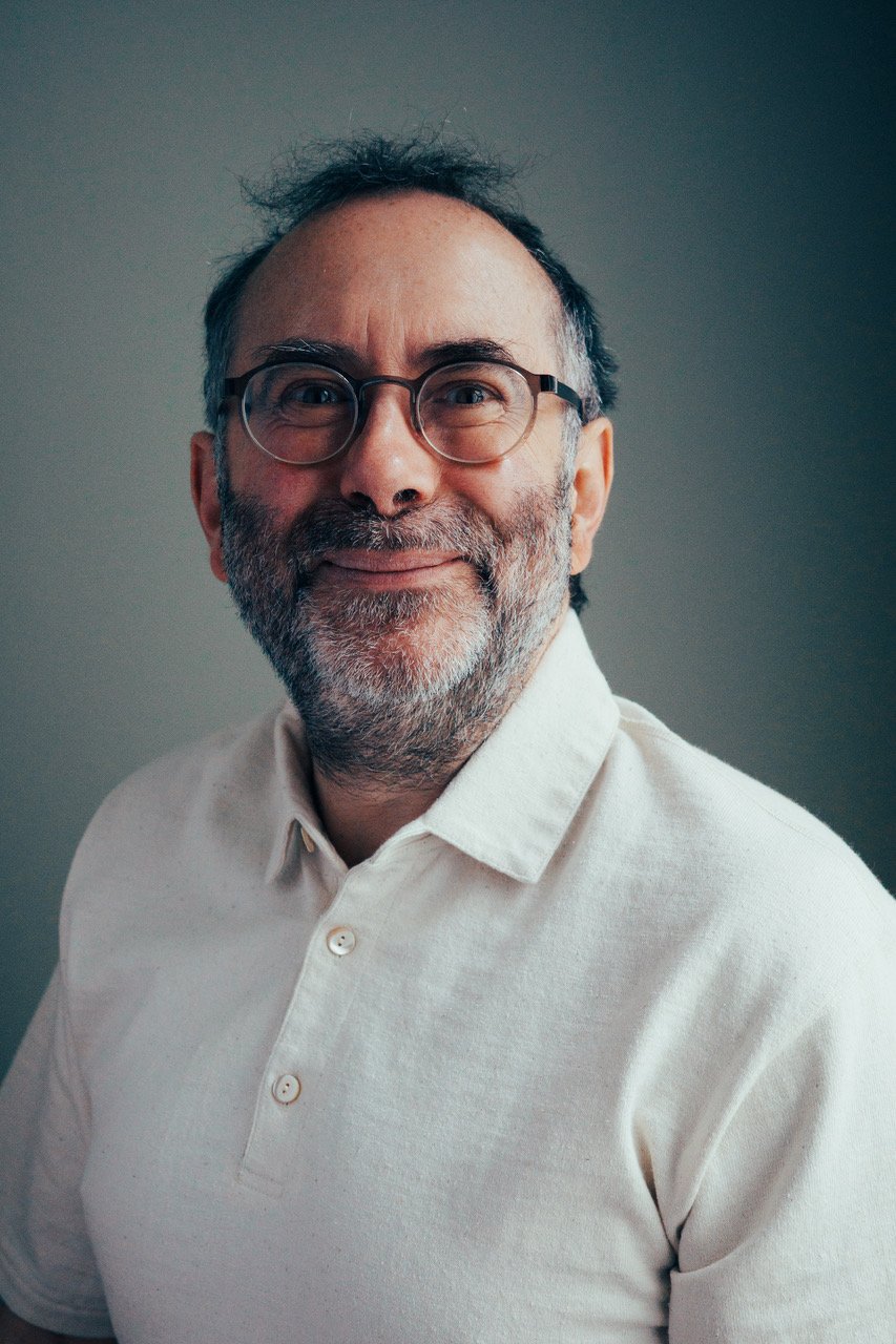 A middle-aged man with glasses and a beard, wearing a white collared shirt, smiling at the camera with a neutral background.