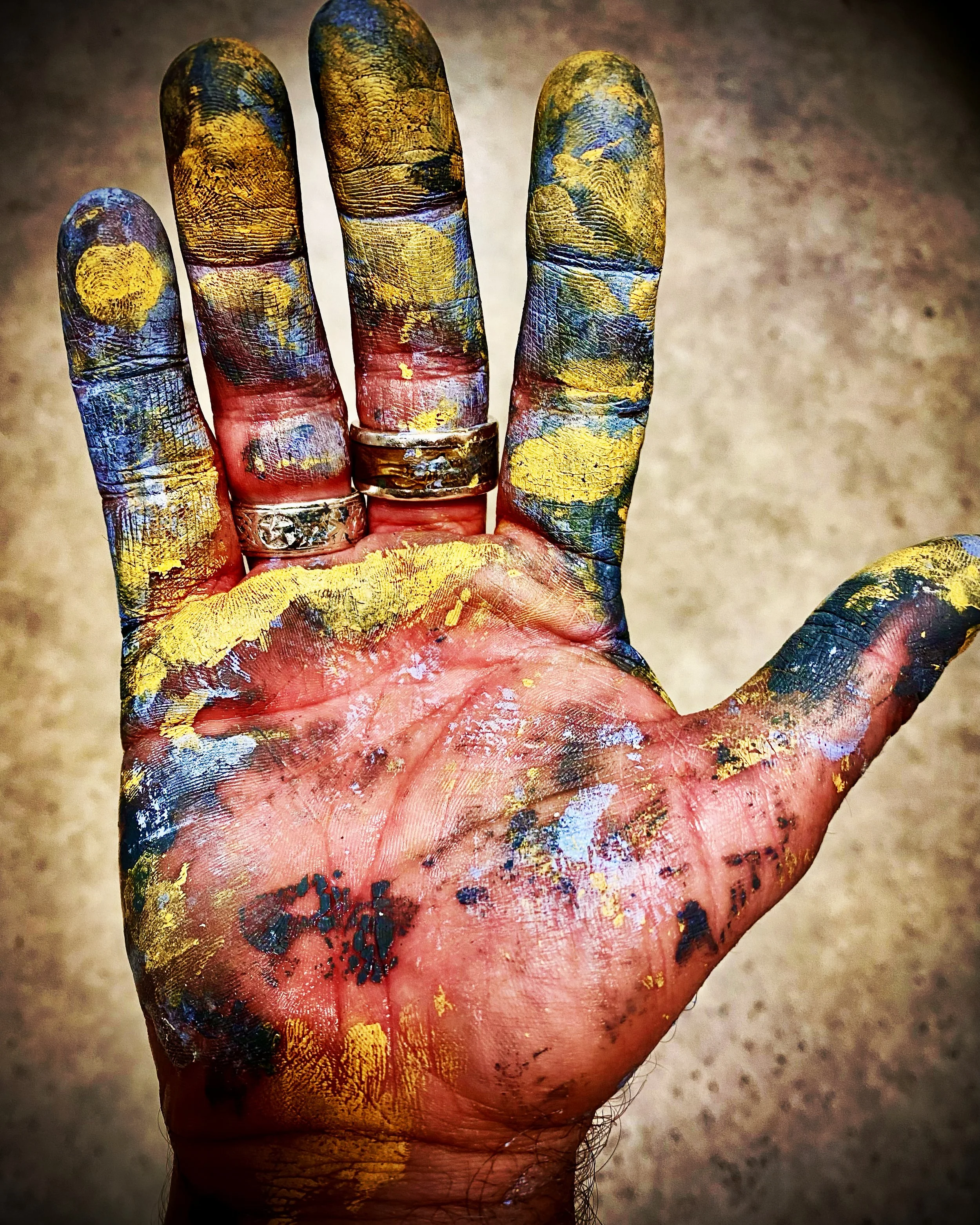 A person's hand covered in colorful paint splatters, with rings on the fingers, against a neutral background.