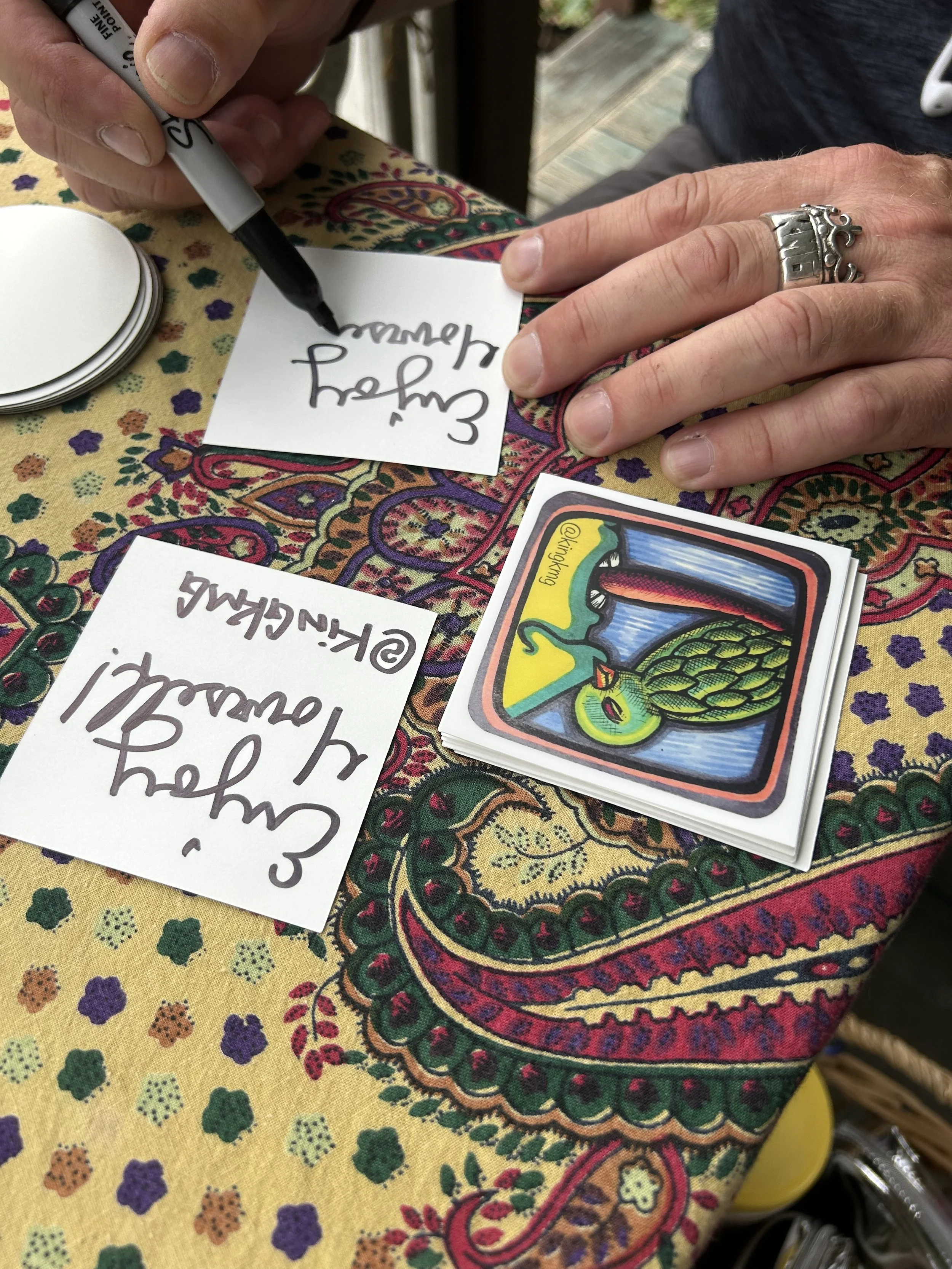 A person with a silver ring on their finger is signing a white card with a black marker. The table has a colorful, patterned cloth and a stack of cards featuring a colorful illustration of a dragon-style creature with a green body and red tongue. The person's other hand is resting on the table, near a handwritten note that says "Enjoy your reading."