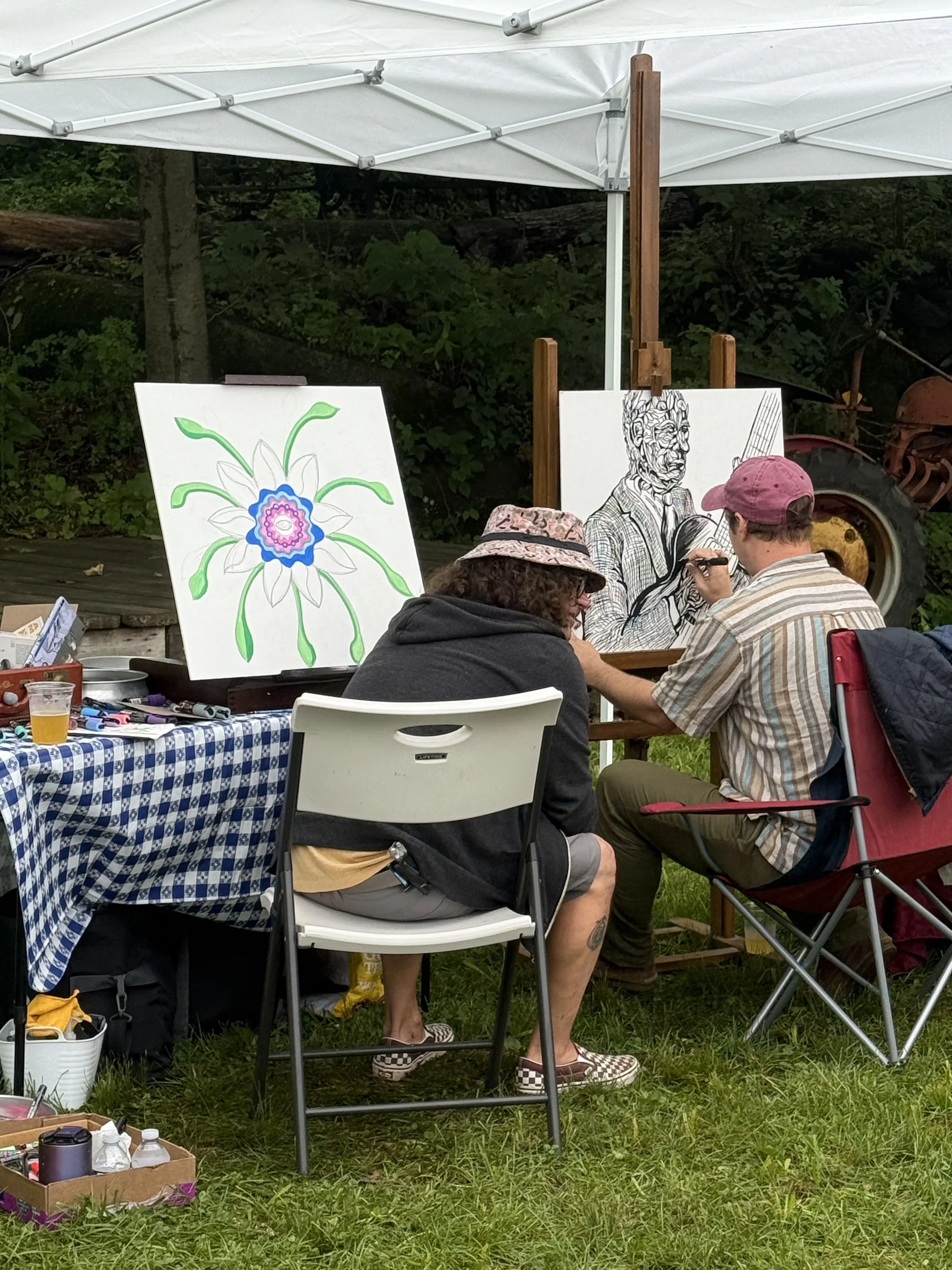 Two artists painting at an outdoor event under a white canopy. One artist is drawing a detailed black-and-white portrait of a man with a bow and arrow on a canvas, while the other is working on a colorful, abstract flower painting with green, white, blue, and pink colors. There are art supplies and drinks on a checkered tablecloth on the table in front of them, with green grass and trees in the background.