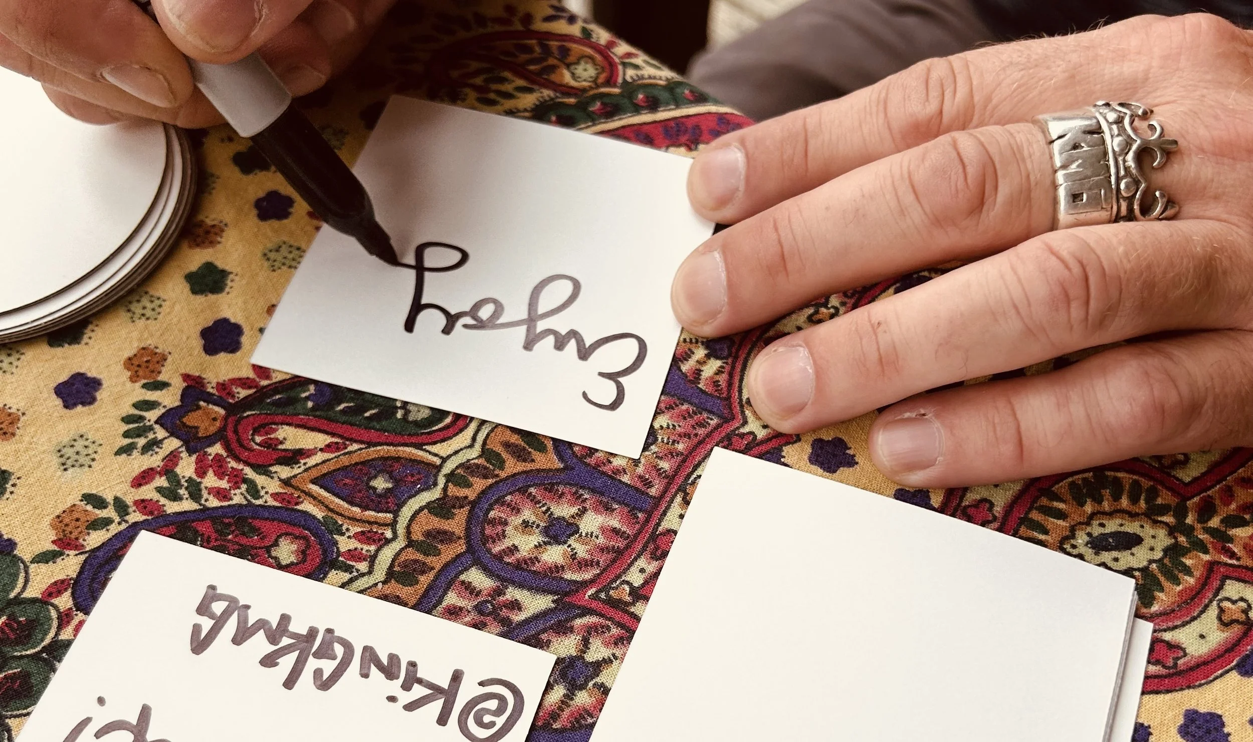 A person writing on a white card with a black marker, while wearing a silver crown ring on their middle finger and a decorative silver ring on their index finger. The person is sitting at a table with a colorful, intricate patterned tablecloth and stacks of other cards.