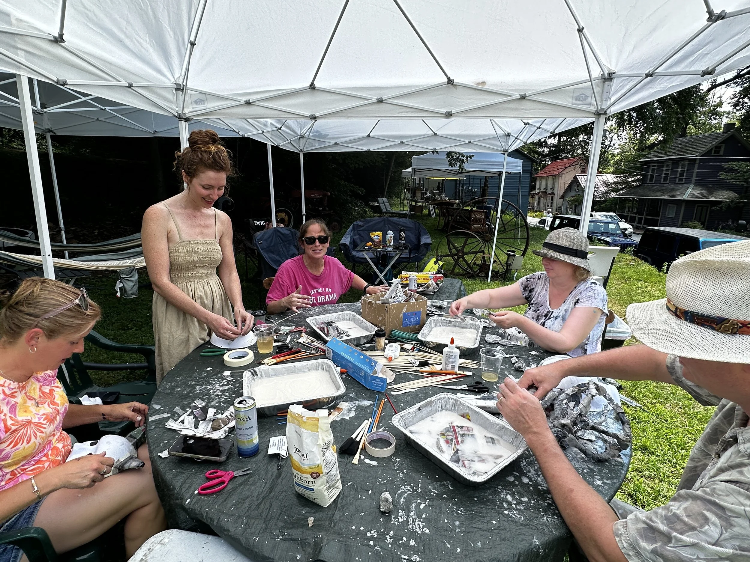 A group of five women sitting and standing around a table outdoors, engaged in a craft activity under a white canopy. The table is covered with craft supplies, including paint, brushes, and foil, on a grassy yard with houses and parked cars in the background.