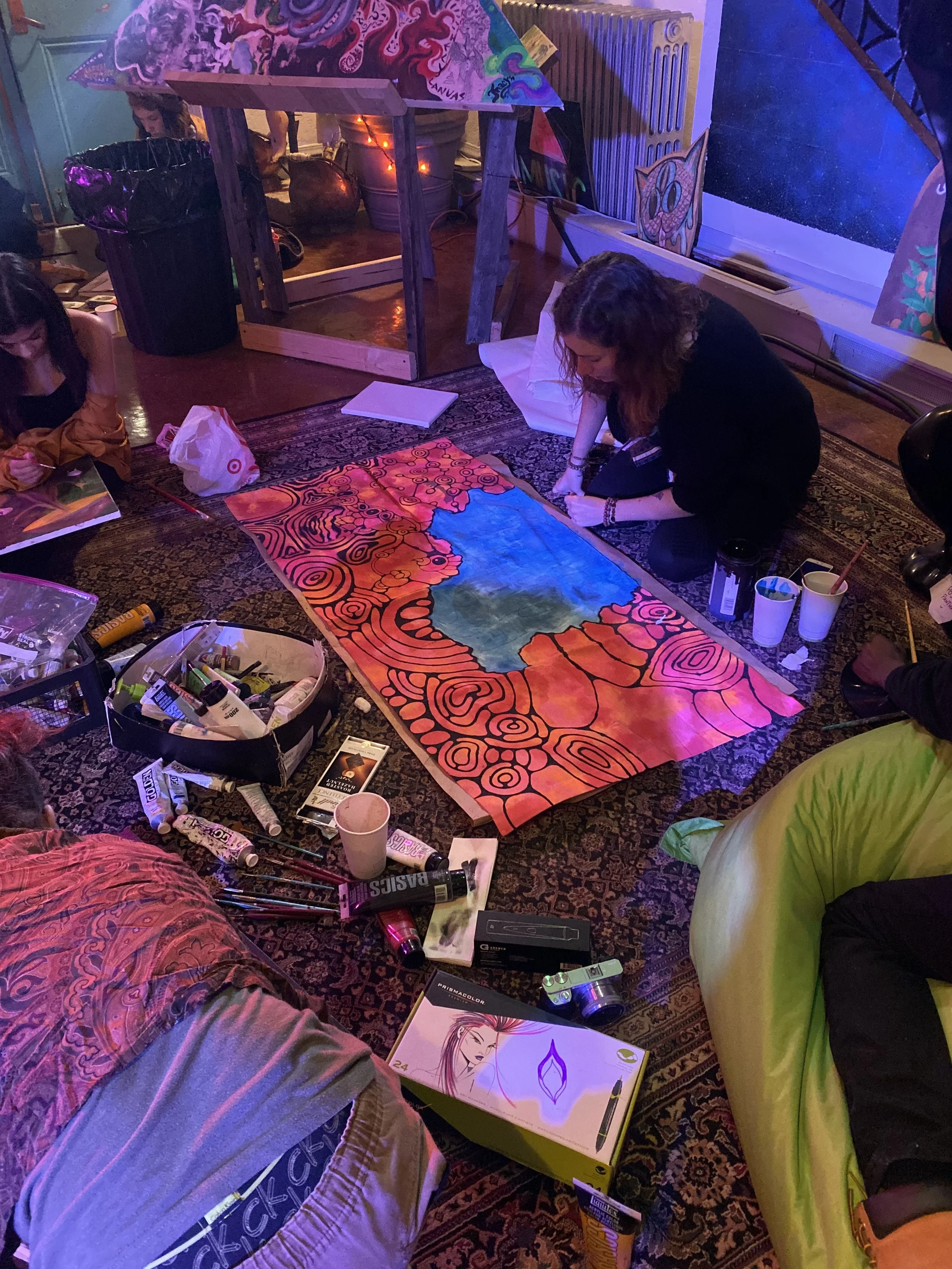 People creating a colorful abstract painting on paper, surrounded by art supplies in an indoor space with a patterned rug and colorful decorations.