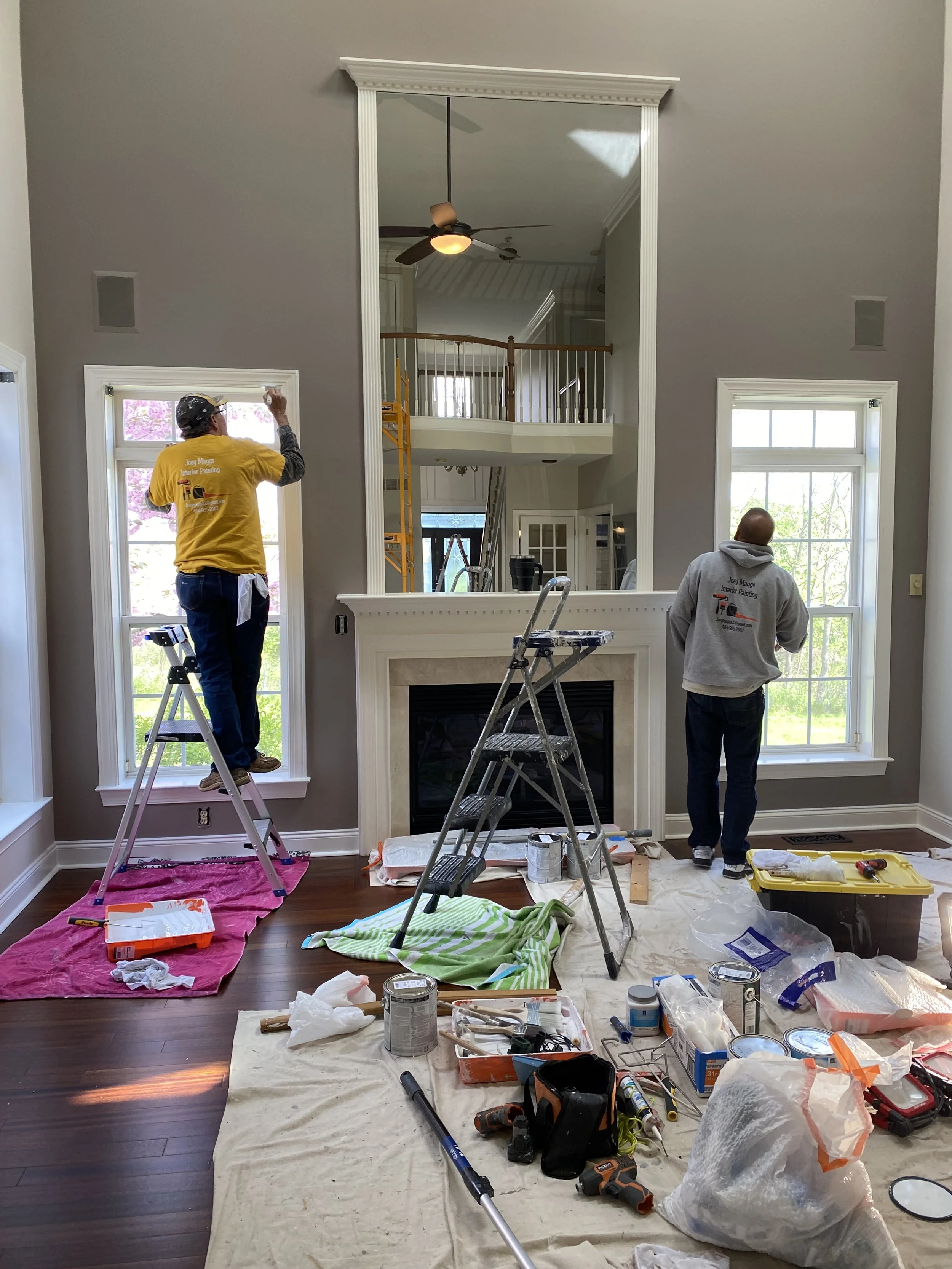 Two workers painting window frames in a living room during renovation, with tools and supplies scattered on the floor.