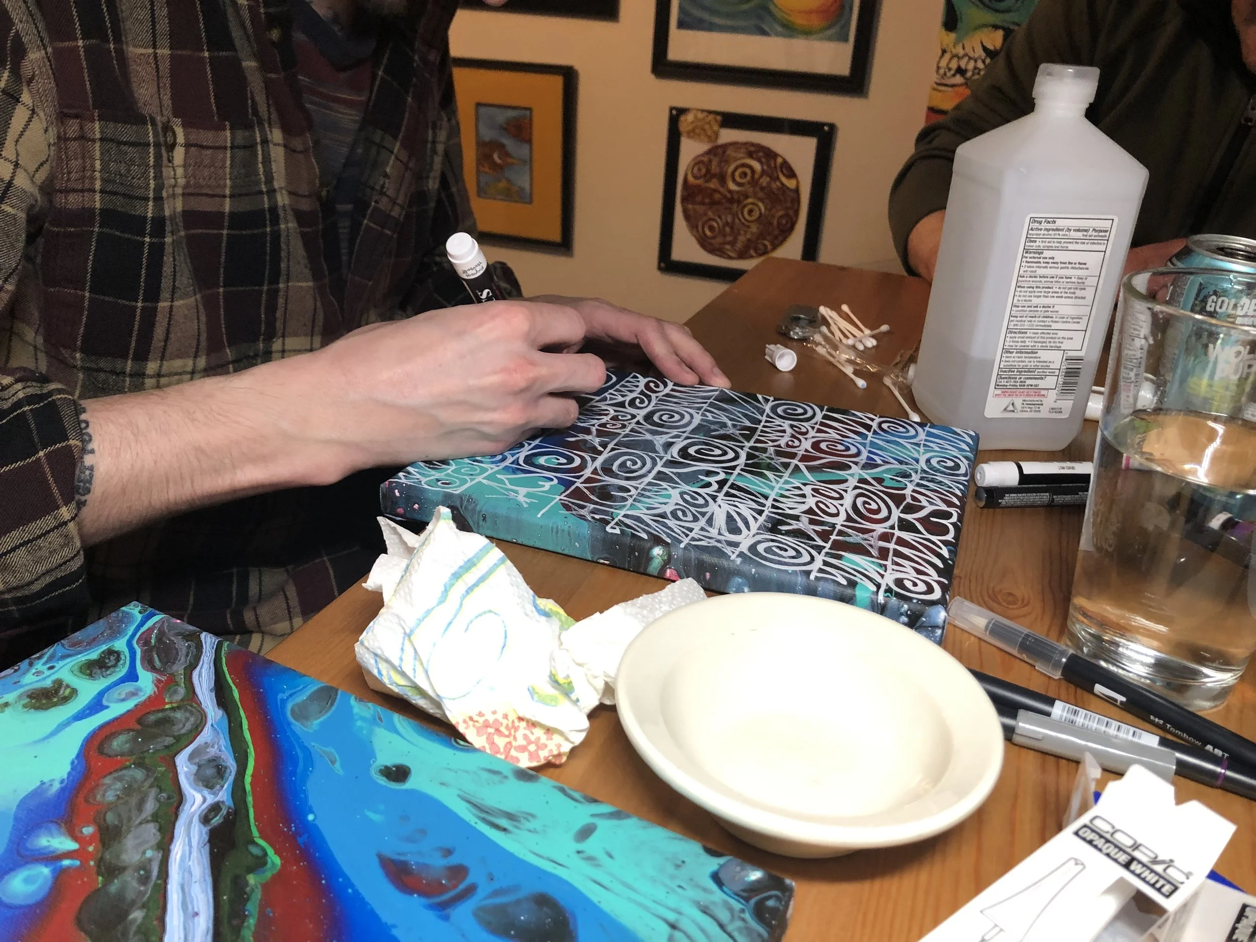 People working on abstract paintings with paint, markers, and paper towels on a wooden table. Artwork featuring swirling patterns and vibrant colors is visible.