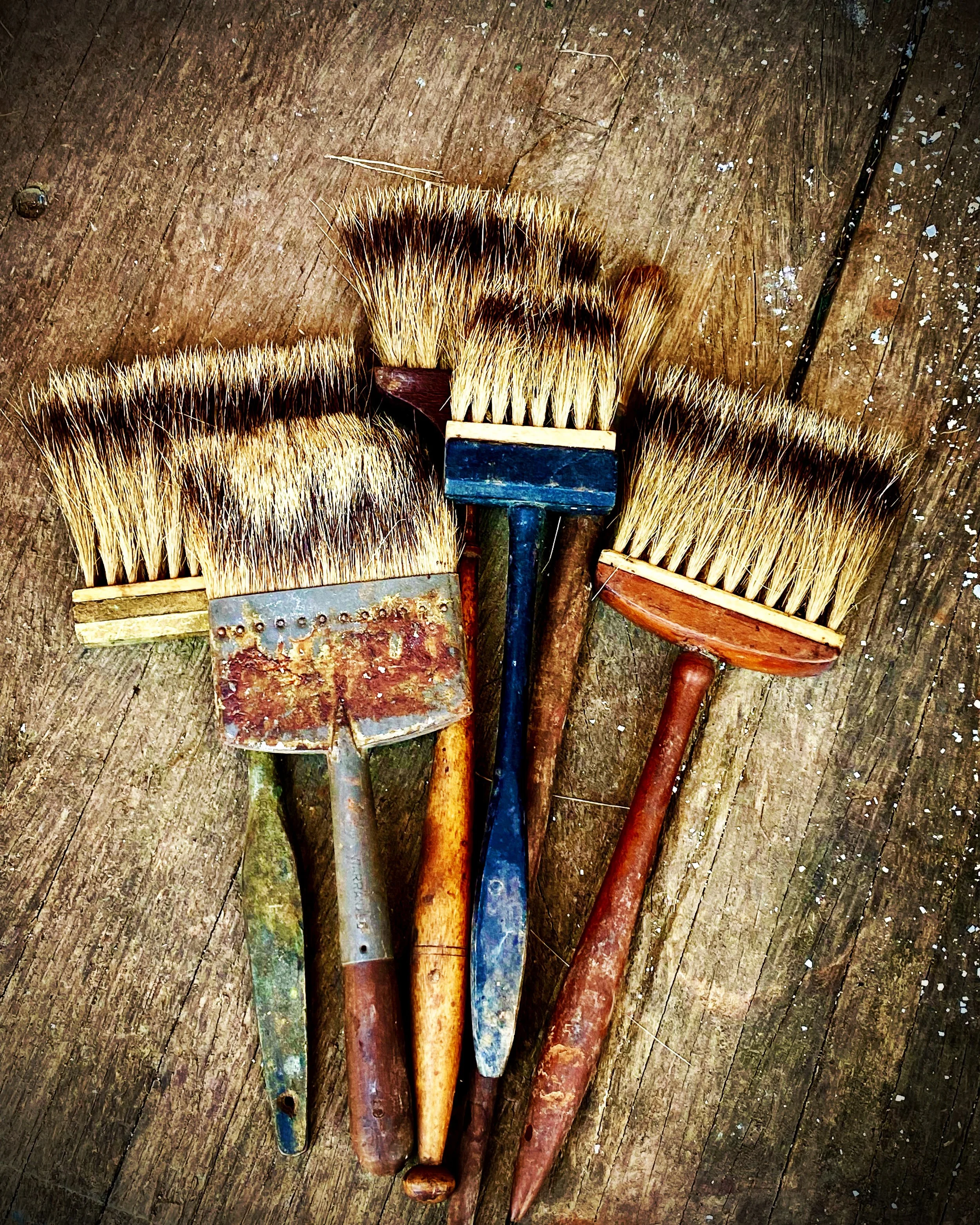 Four old wire brushes with wooden and metal handles spread out on a weathered wooden surface.