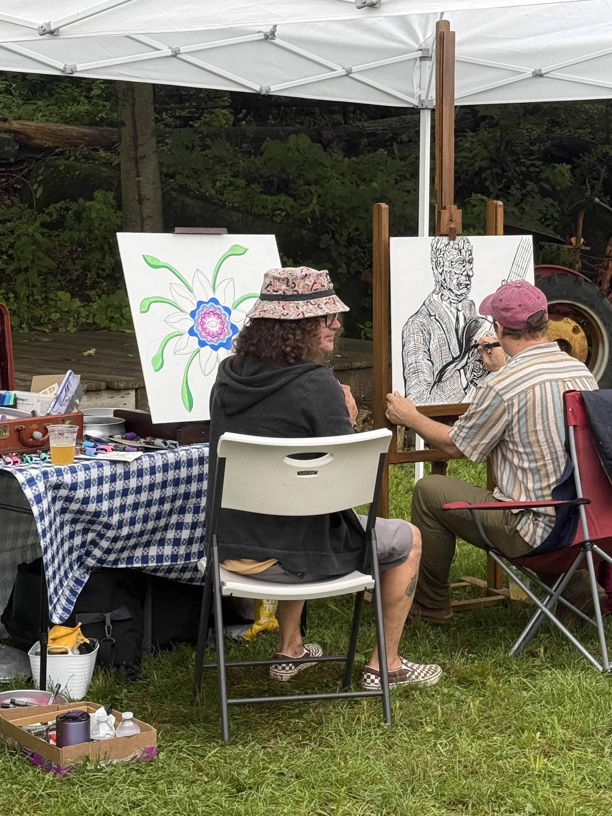 Two men are painting outdoors under a white canopy. One man is seated, wearing a pink cap and striped shirt, and is drawing a sketch of a person on a canvas. The other man is sitting backwards in a white folding chair, wearing a hat, hoodie, and shorts, looking at the painting. A table with art supplies and a drink is nearby, with trees and a tire in the background.