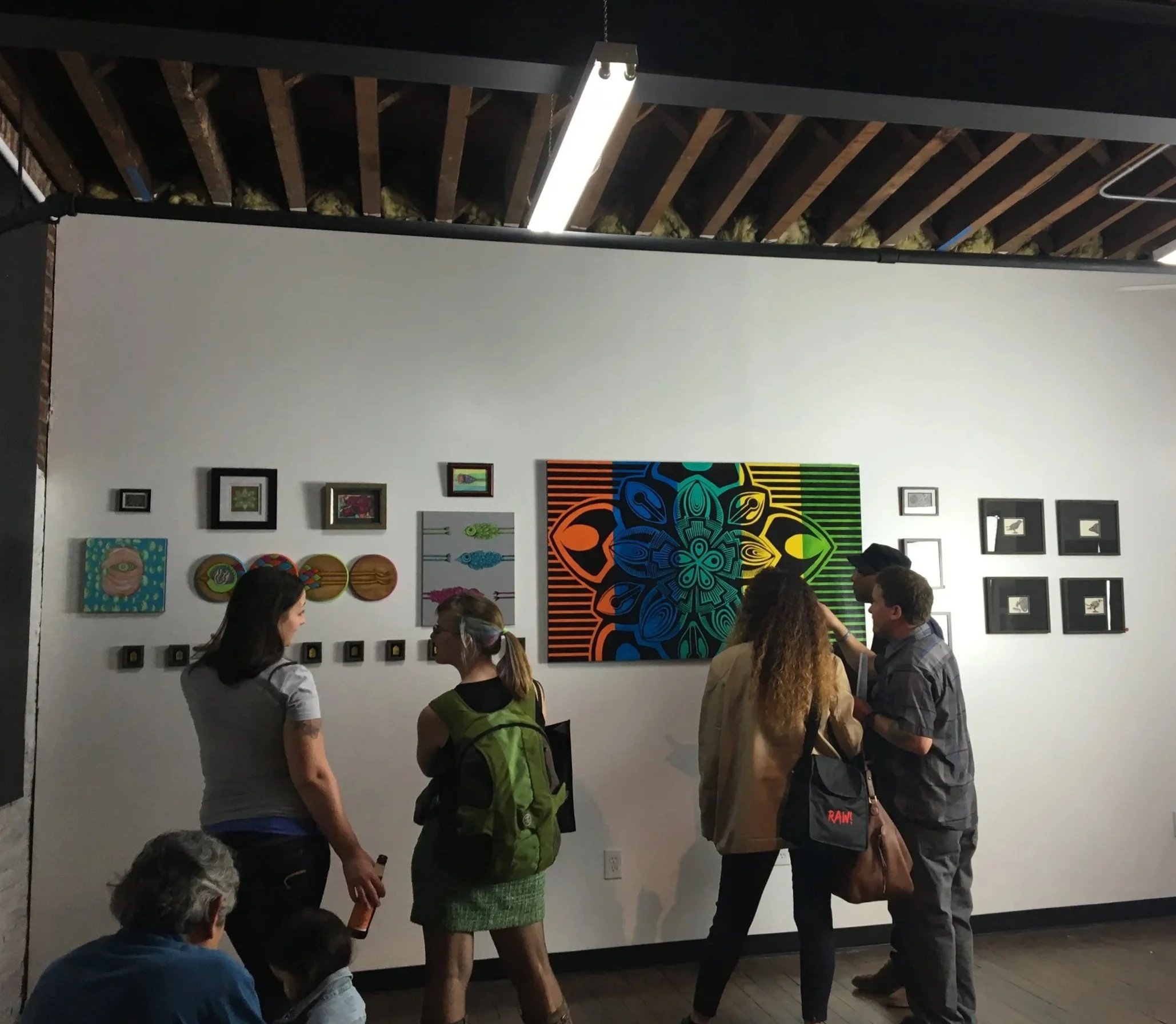 People observing various colorful artworks, including paintings and framed pictures, on a white gallery wall in an art exhibition.