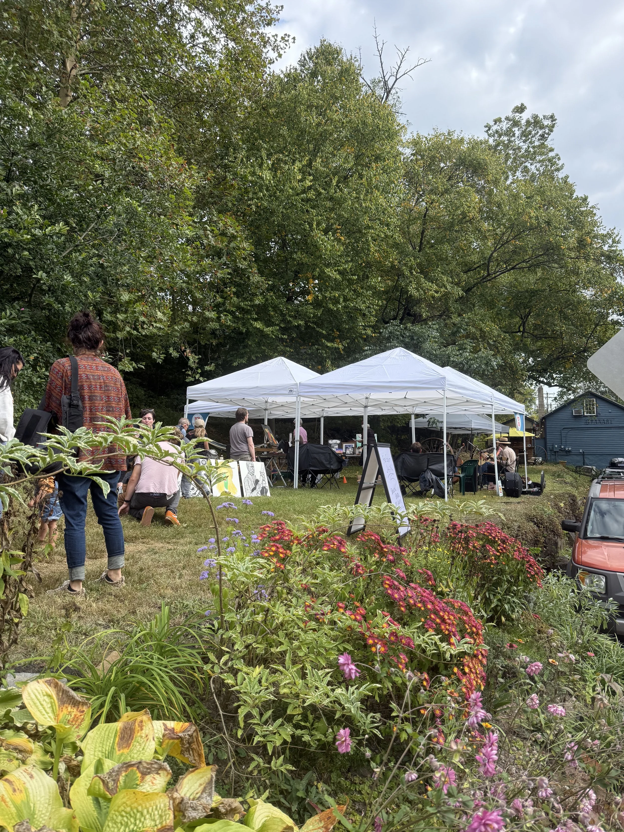 A daytime outdoor art fair with white tents set up on a grassy area surrounded by trees and colorful flowers. People are walking, browsing, and sitting near the tents.