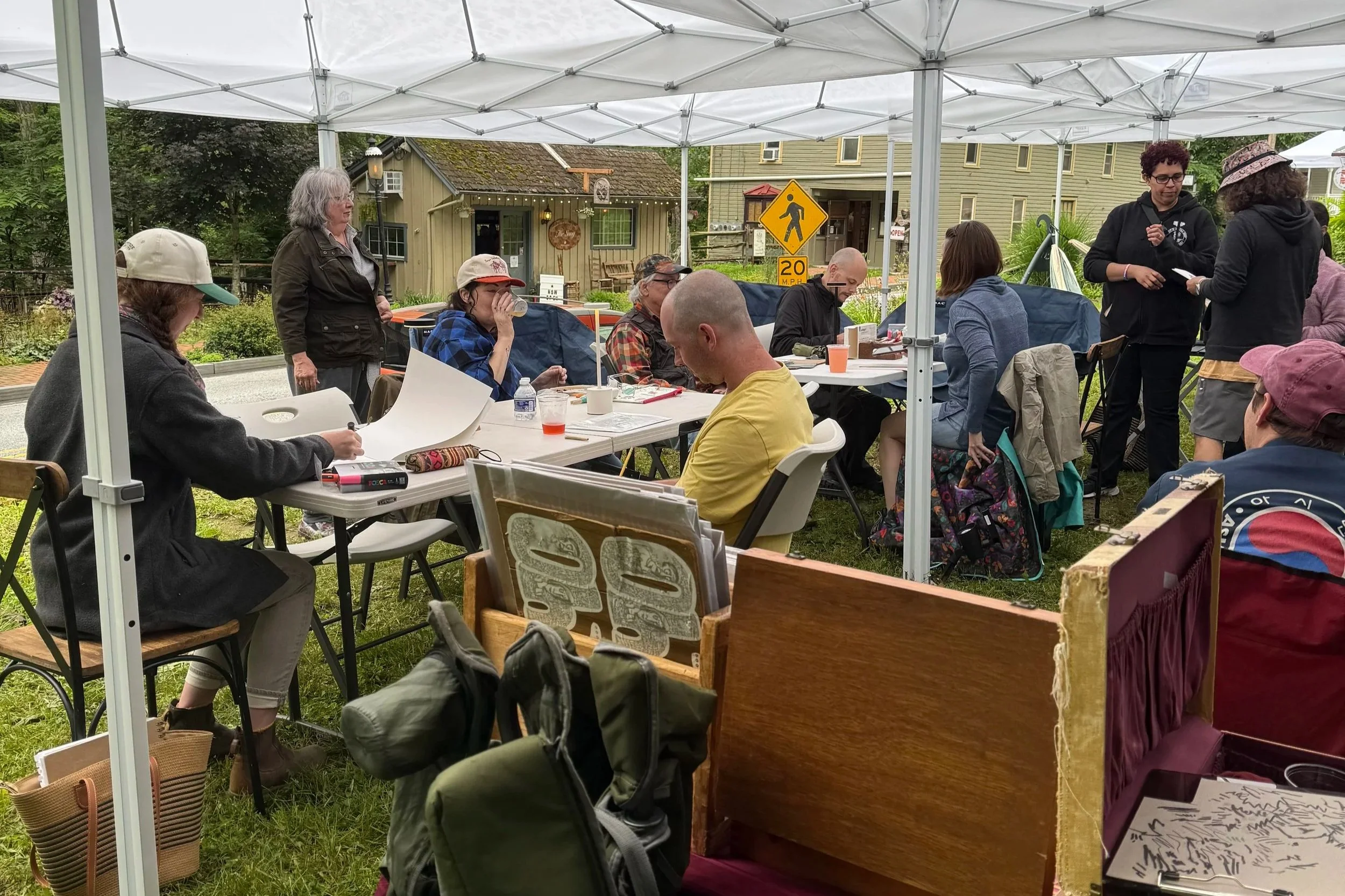 People gathered under a white canopy tent outdoors, sitting at tables with papers, drinks, and backpacks, with a yellow pedestrian crossing sign and a small house in the background.