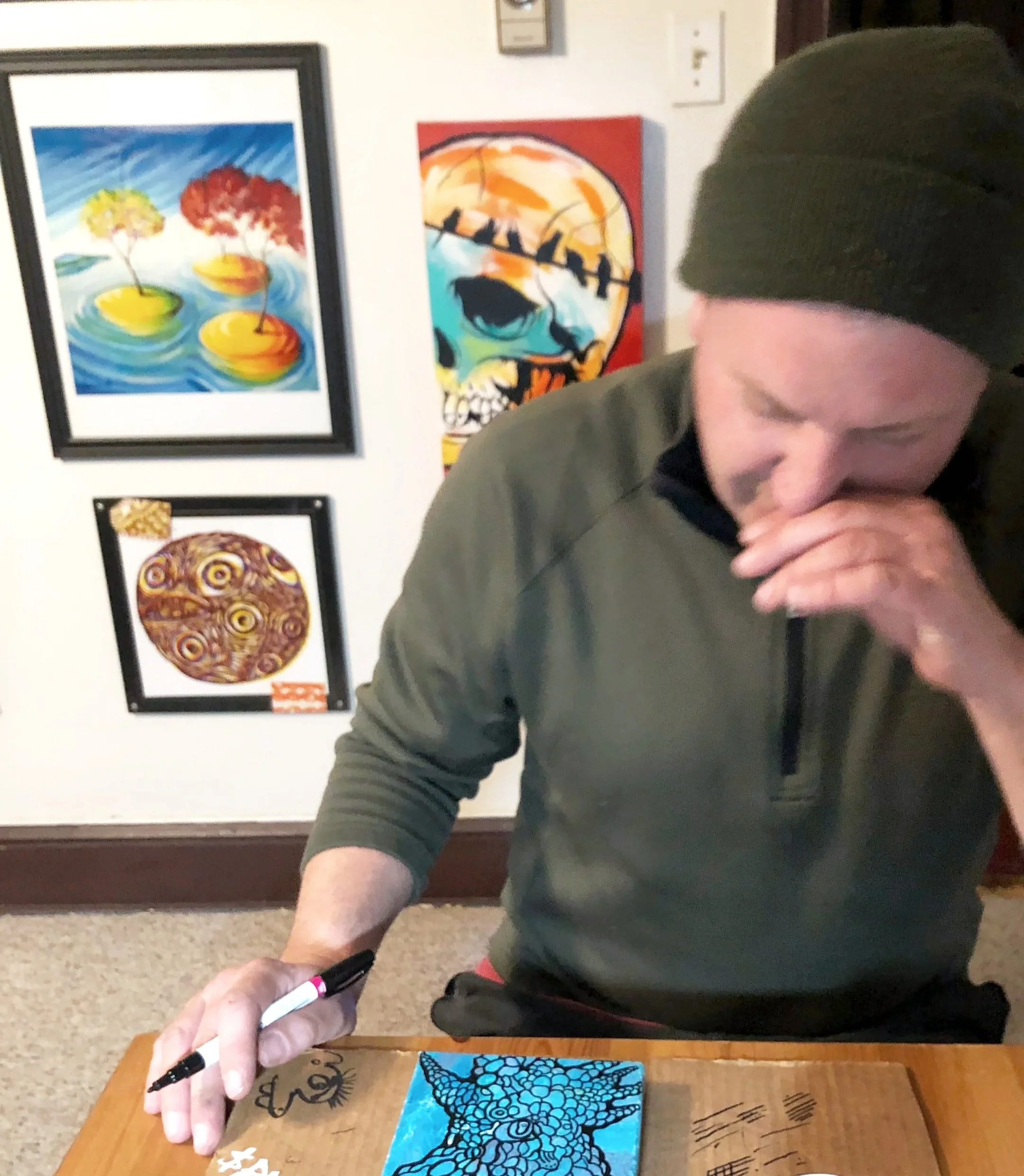 A person wearing a dark beanie and green shirt is sitting at a wooden table, holding a black marker and a blue artwork featuring a black web-like pattern. The person is covering their mouth with one hand. Behind them are four colorful paintings on a white wall, including a tree scene with water, a skull with other designs, and abstract circular patterns.