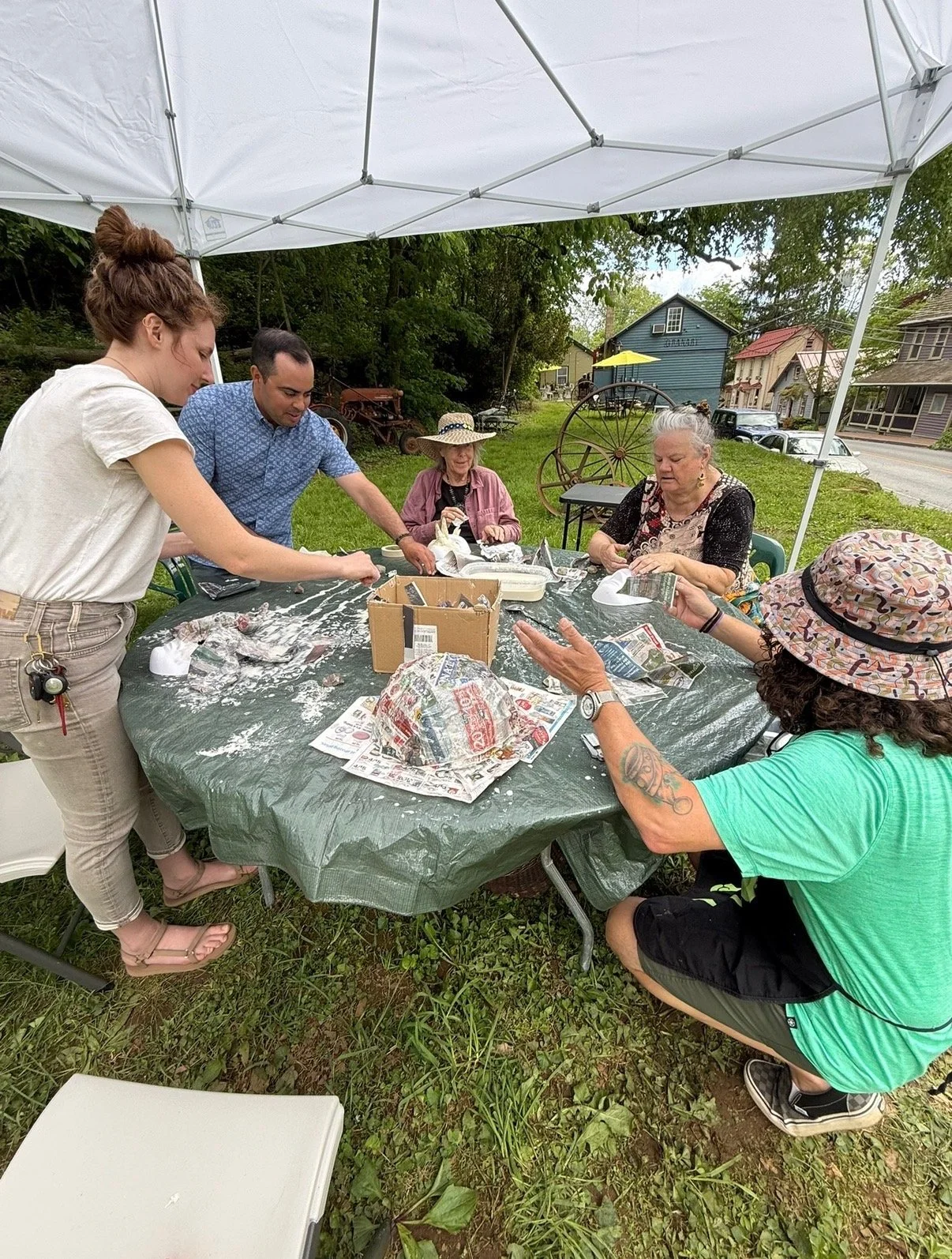A group of six people gathered around a table outdoors under a canopy, working on a craft project with plaster and newspaper, with a grassy yard and houses in the background.