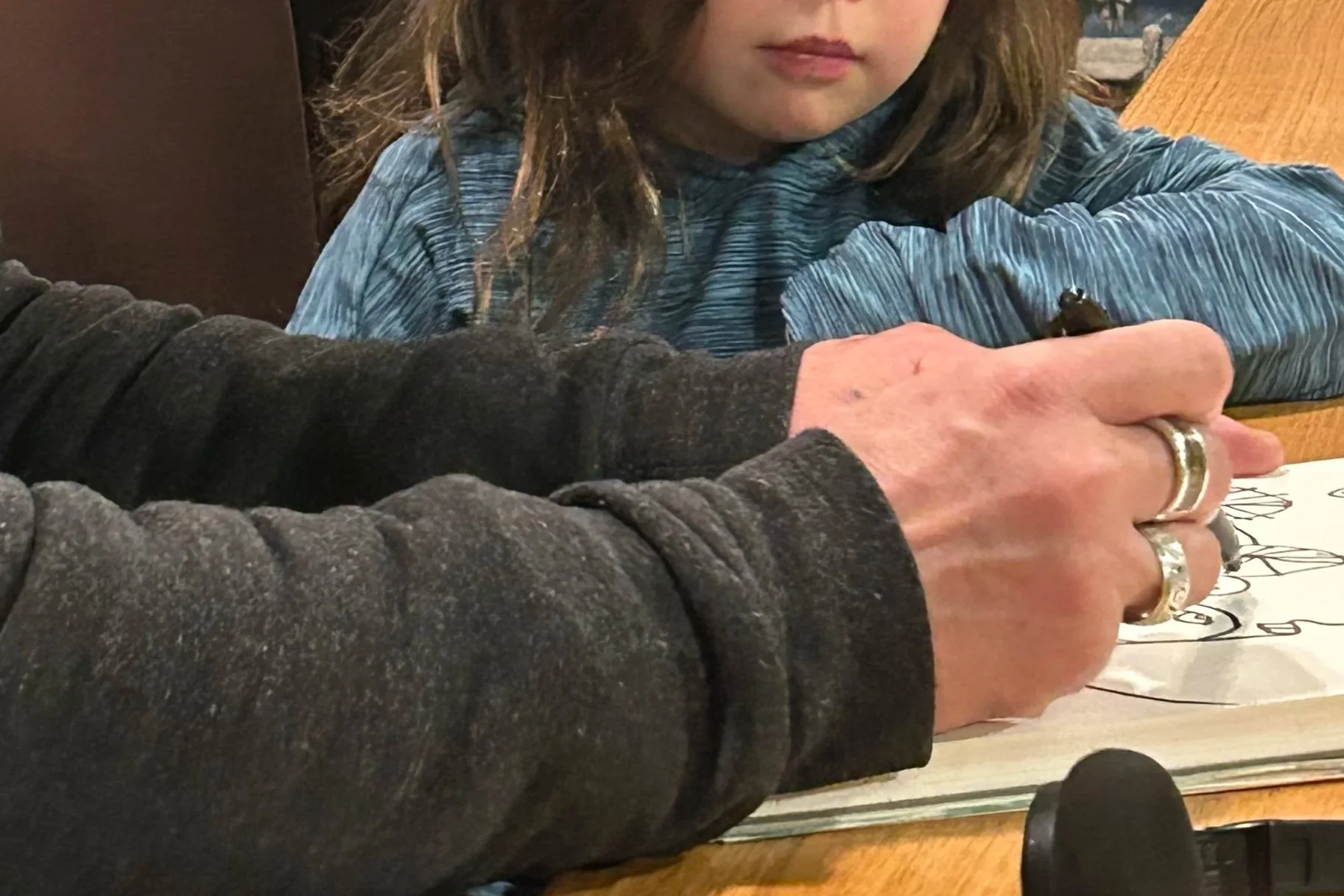 A child with long hair and a blue shirt sitting at a table. An adult, wearing a black sweater and silver rings, is pointing or drawing on a piece of paper on the table, which has a black and white design. The adult's hand and arm are in the foreground, partially obscuring the child's face.