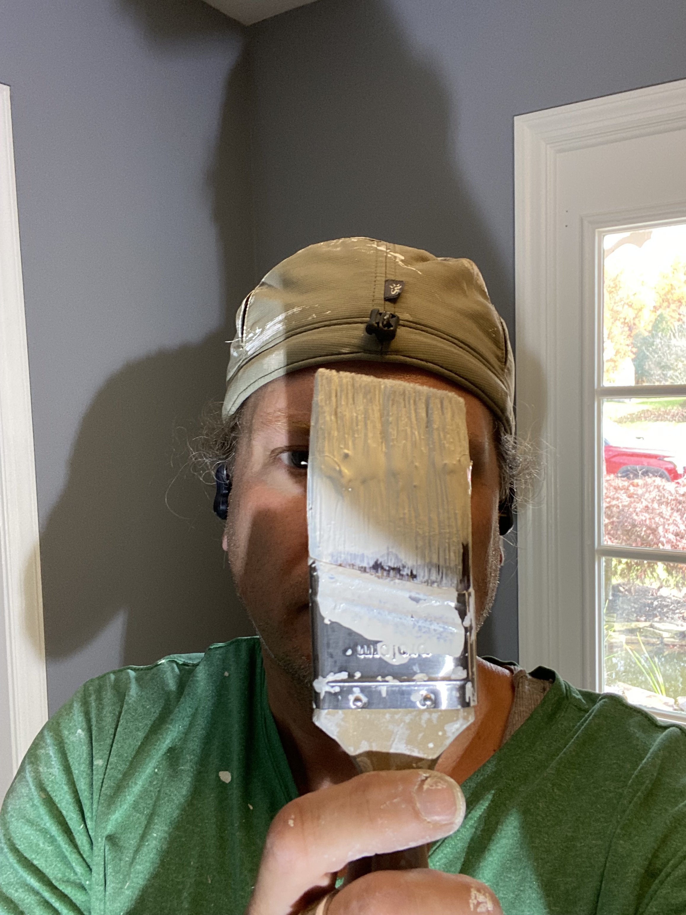 Person holding a paintbrush in front of their face, wearing a beige cap and green shirt, with a window in the background.