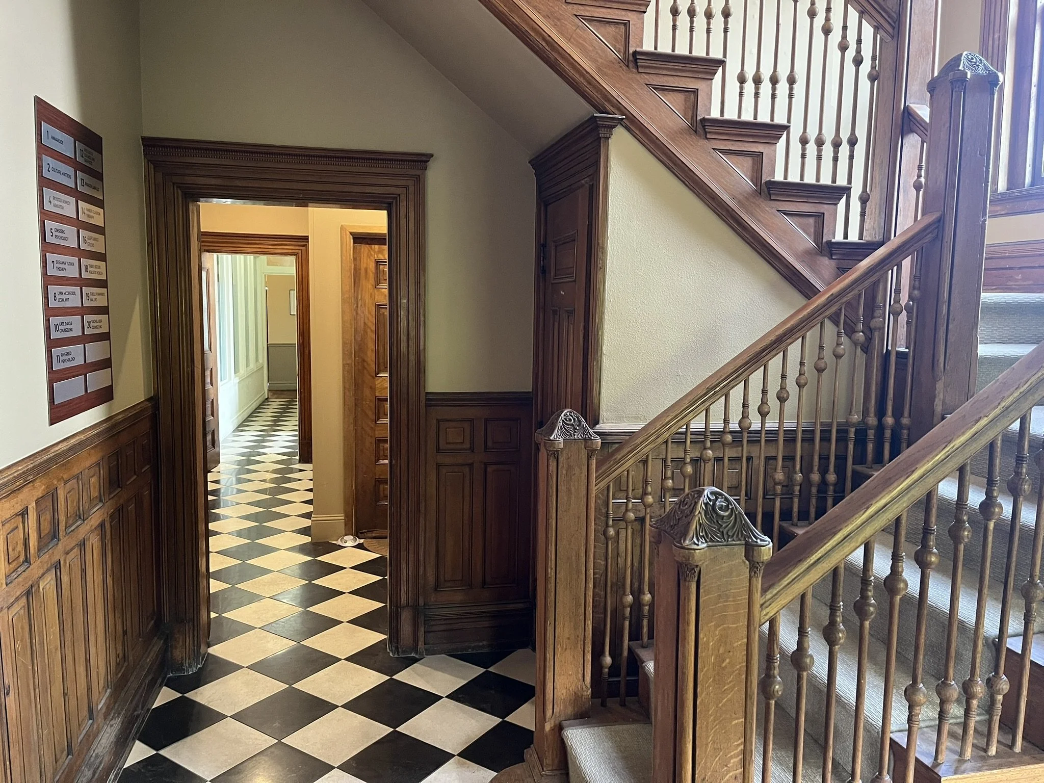 Interior view of a building with wooden wainscoting, staircase with wooden spindles, and a black-and-white checkered tile floor.